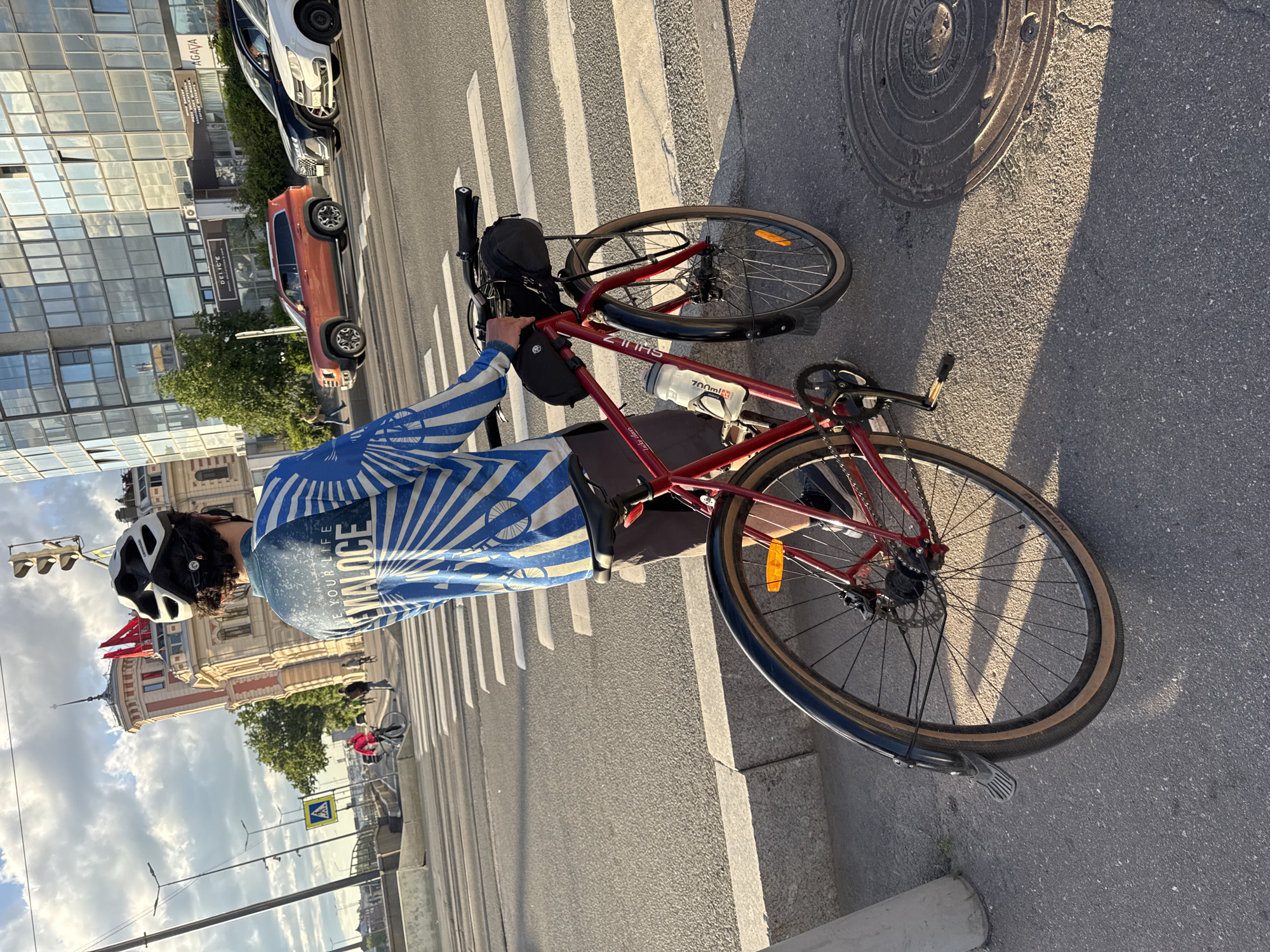 The image captures a cyclist paused at a crosswalk in Saint Petersburg, Russia, with the city's architecture serving as a backdrop. The cyclist, wearing a blue and white long-sleeved shirt with a sunburst pattern and a white helmet, is standing next to a red bicycle. The bicycle has a black saddlebag on the handlebars and a water bottle attached to the frame. The cyclist's back is mostly to the camera, and they appear to be looking towards the street.
The background features a modern building with a glass facade, reflecting the cloudy sky. To the left, a historic building with red accents and a tower can be seen. Several cars are parked and in motion on the street, including an orange SUV and a white car. The crosswalk is clearly marked on the asphalt road. The overall scene suggests a daytime setting with diffused sunlight, possibly late afternoon given the long shadows. The presence of the distinctive architecture and the street scene firmly places the location in Saint Petersburg.