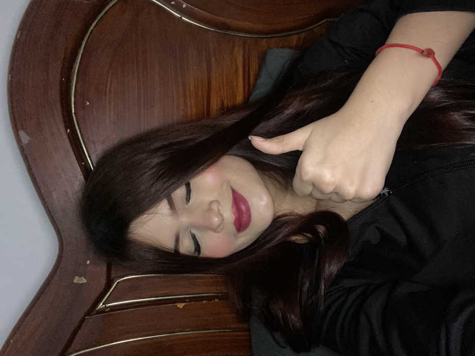 A woman with long, dark hair poses on a wooden headboard. She is reclined, facing the camera with closed eyes and a faint smile. Her lips are painted a deep red, and her cheeks have a rosy blush. She wears a black top, possibly a jacket with a visible zipper. Her right hand gives a thumbs-up, adorned with a red string bracelet. The background features an ornate, dark wood headboard, adding warmth to the image.