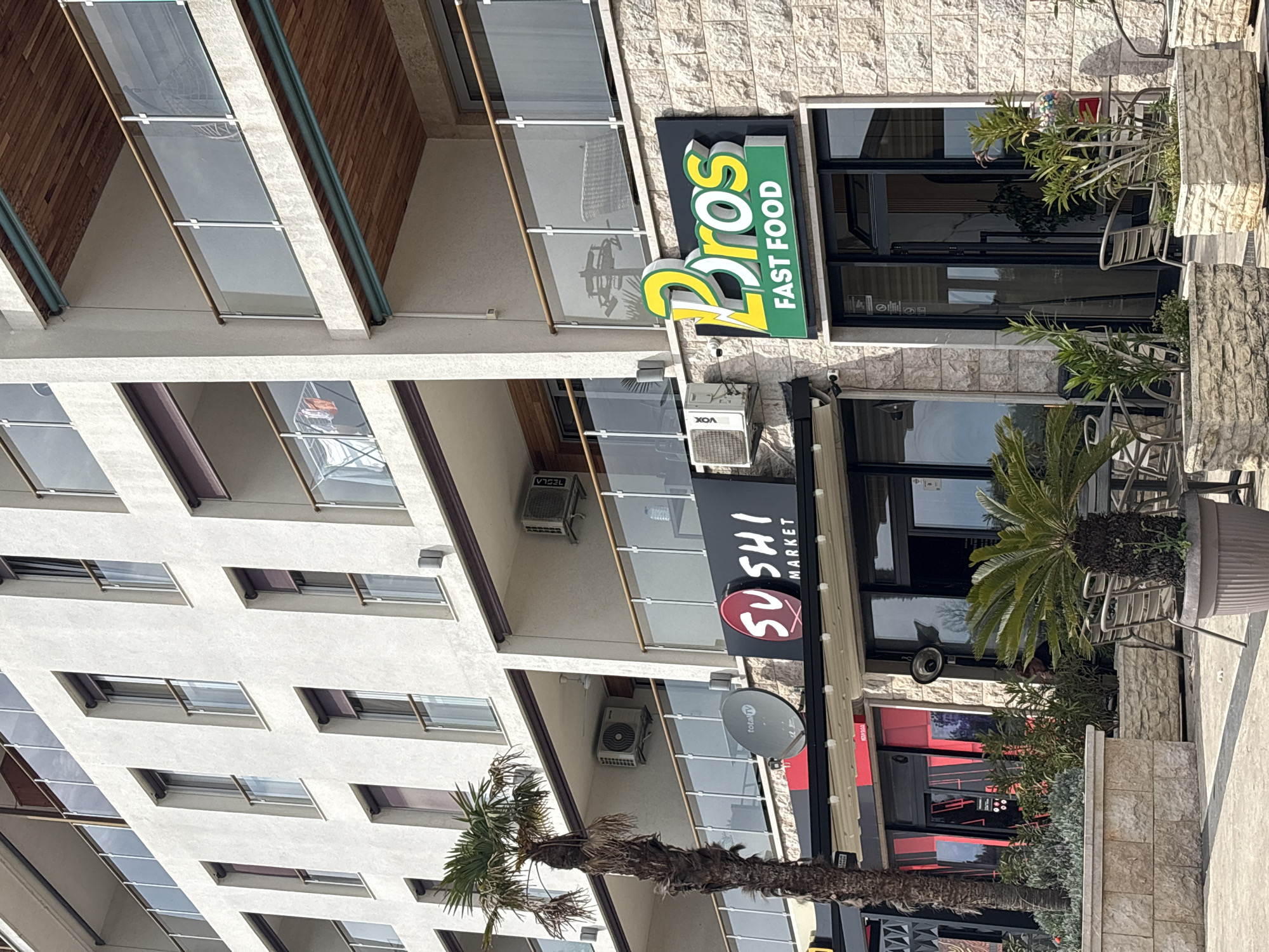 The image shows the front of a building in Bar, Montenegro, featuring two restaurants: "2 Bros Fast Food" and "Sushi Market." The building is multi-storied with white facade and several windows. The restaurants have outdoor seating areas with tables, chairs, and planters. The "2 Bros Fast Food" sign is green and yellow, while the "Sushi Market" sign is black and red. A palm tree is located near the "Sushi Market" entrance. Air conditioning units are mounted on the building's exterior. There is also satellite dish. Pedestrian sidewalk is visible in the foreground. The photograph is taken during daylight hours with natural lighting.