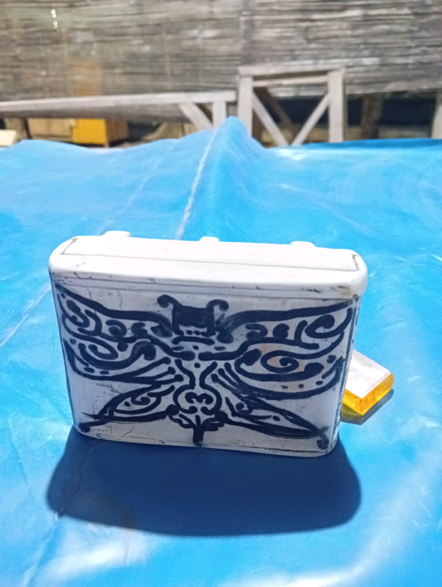 A close-up, eye-level shot shows a rectangular, white container with intricate black designs, possibly depicting a stylized butterfly or dragon, on its front surface. The container appears to be made of ceramic or painted plastic and has a slightly weathered appearance. It rests on a bright blue, crinkled surface, which could be a tarp or a tablecloth. To the right and slightly behind the container, a small, translucent yellow object, possibly a piece of hard candy or resin, is visible. The background is blurred but suggests an outdoor setting with wooden structures and possibly a corrugated metal roof. The lighting suggests it is daytime, with a strong shadow cast by the container on the blue surface. The location is Pucakwangi, Indonesia.