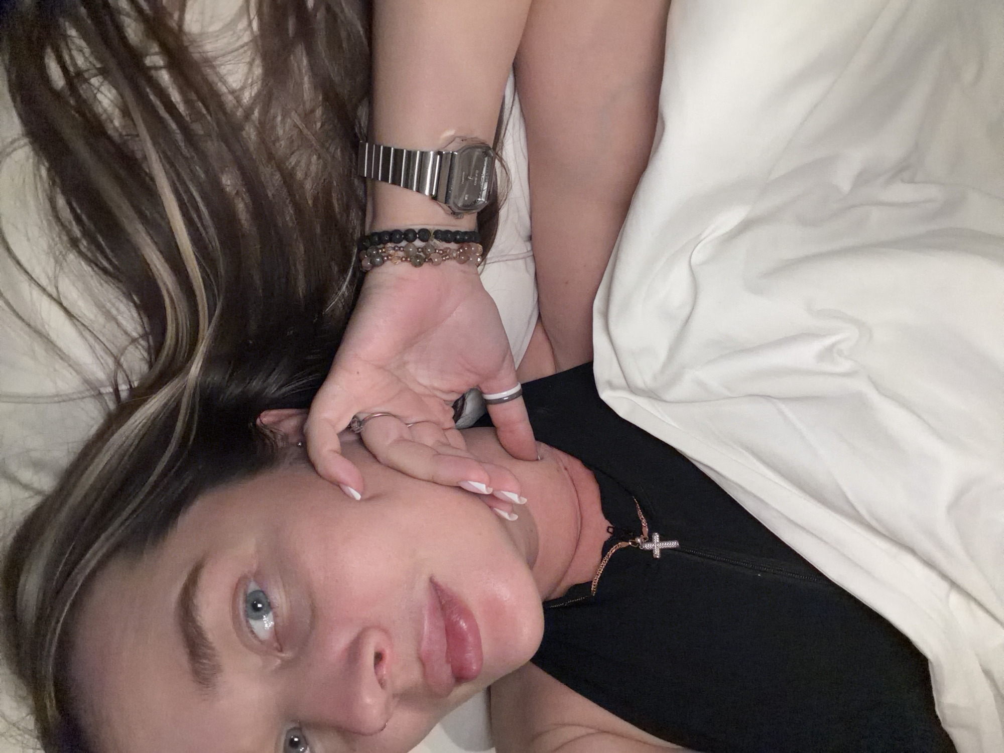 A woman with light skin, brown hair with blonde highlights, and blue eyes is lying on a white bed, possibly in a hotel room. She is wearing a silver wristwatch and several bracelets on her left wrist, along with rings on her fingers. She is also wearing a black top with a silver cross necklace. The location in the city of Moscow, Russia cannot be determined from the image.