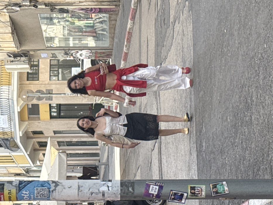 The image displays two young women walking on an asphalt street in Tel Aviv-Yafo, Israel, under bright daylight.
The woman on the left wears a white, strappy corset-style top, a black lace midi skirt, and gold flat shoes. She has dark hair, a dark choker necklace, and carries a light-colored tote bag on her left shoulder. Her mouth is slightly open, and she looks to her right.
The woman on the right wears a red sleeveless crop top with white text (partially visible as "DIESEL"), white wide-leg trousers, and red flat shoes. A red long-sleeved garment is tied around her waist. She has dark, curly hair, wears a gold necklace, and a white scrunchie on her left wrist. She holds a red shoulder bag and a clear plastic cup containing a light-brown beverage with a straw in her right hand. Her facial expression is neutral with slightly pursed lips.
In the foreground, a grey utility pole on the left is affixed with "Bolt food" advertisements displaying Hebrew text and a QR code, alongside other stickers featuring Hebrew text and images of people. The street pavement is grey asphalt with visible cracks. A red and white striped curb runs along the right side of the street.
The background features buildings typical of Tel Aviv-Yafo. On the left, a light-colored building with an awning is visible, with a person in dark clothing partially seen further down the street. In the center-right, a two-story building displays a yellow upper facade and a lighter lower section with dark doors and windows. A white bicycle is parked near this building. To the far right, a shop window exhibits clothing, and the adjacent wall has graffiti. An outdoor air conditioning unit is mounted on the building facade. A multi-directional street sign pole stands between the buildings. Additional Hebrew text is visible on various signs and posters in the background.