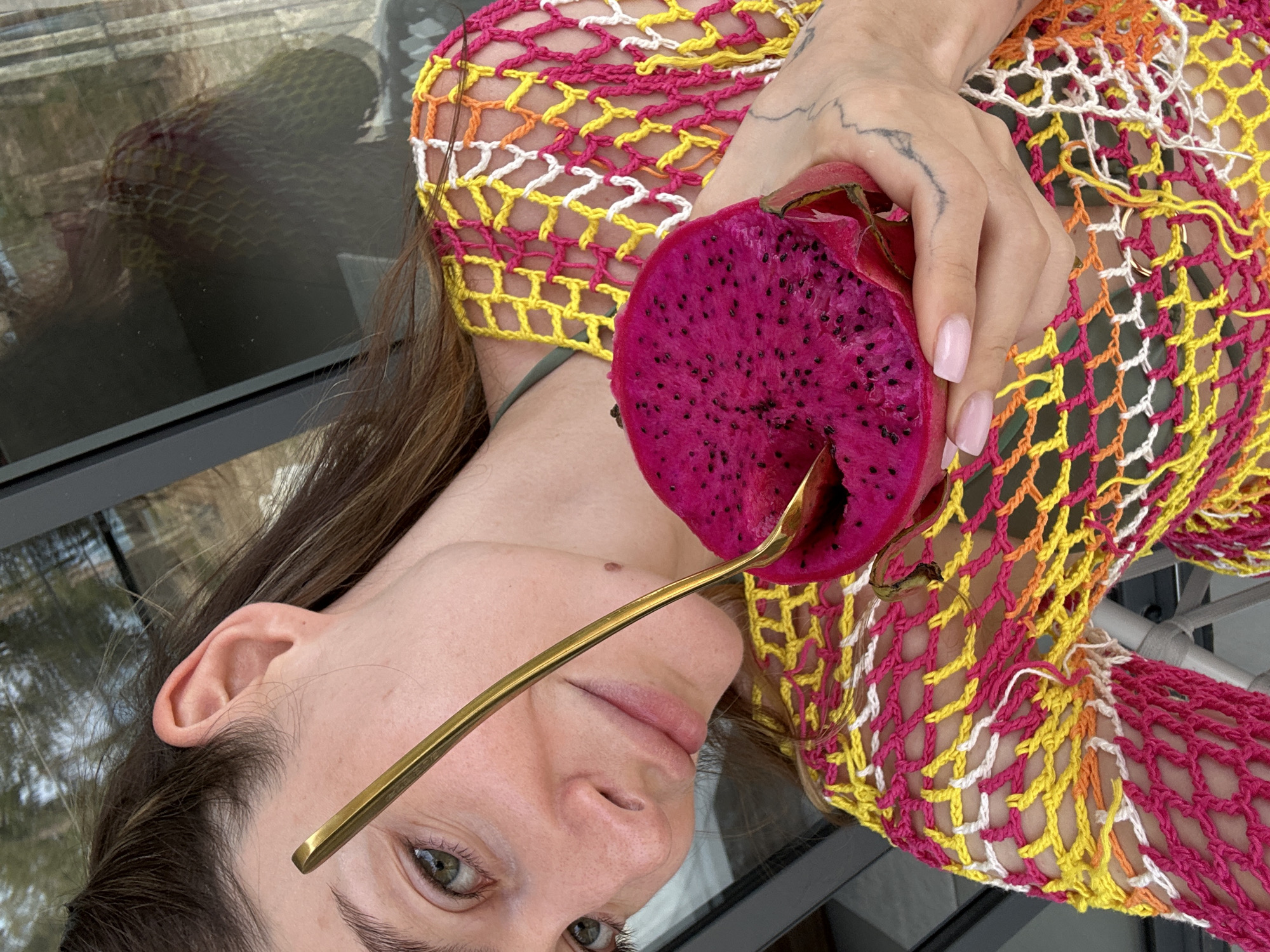A person is shown in a portrait orientation, sideways to the camera. They are holding a halved dragon fruit with a golden spoon inside. The dragon fruit has magenta flesh with small black seeds. The person has fair skin, brown hair, and green eyes. Their fingernails are painted a light pink. They are wearing a colorful crocheted top or headpiece in pink, yellow, orange, and white. The image appears to be taken outdoors on a reflective glass surface. The location is Choeng Thale, Thailand.