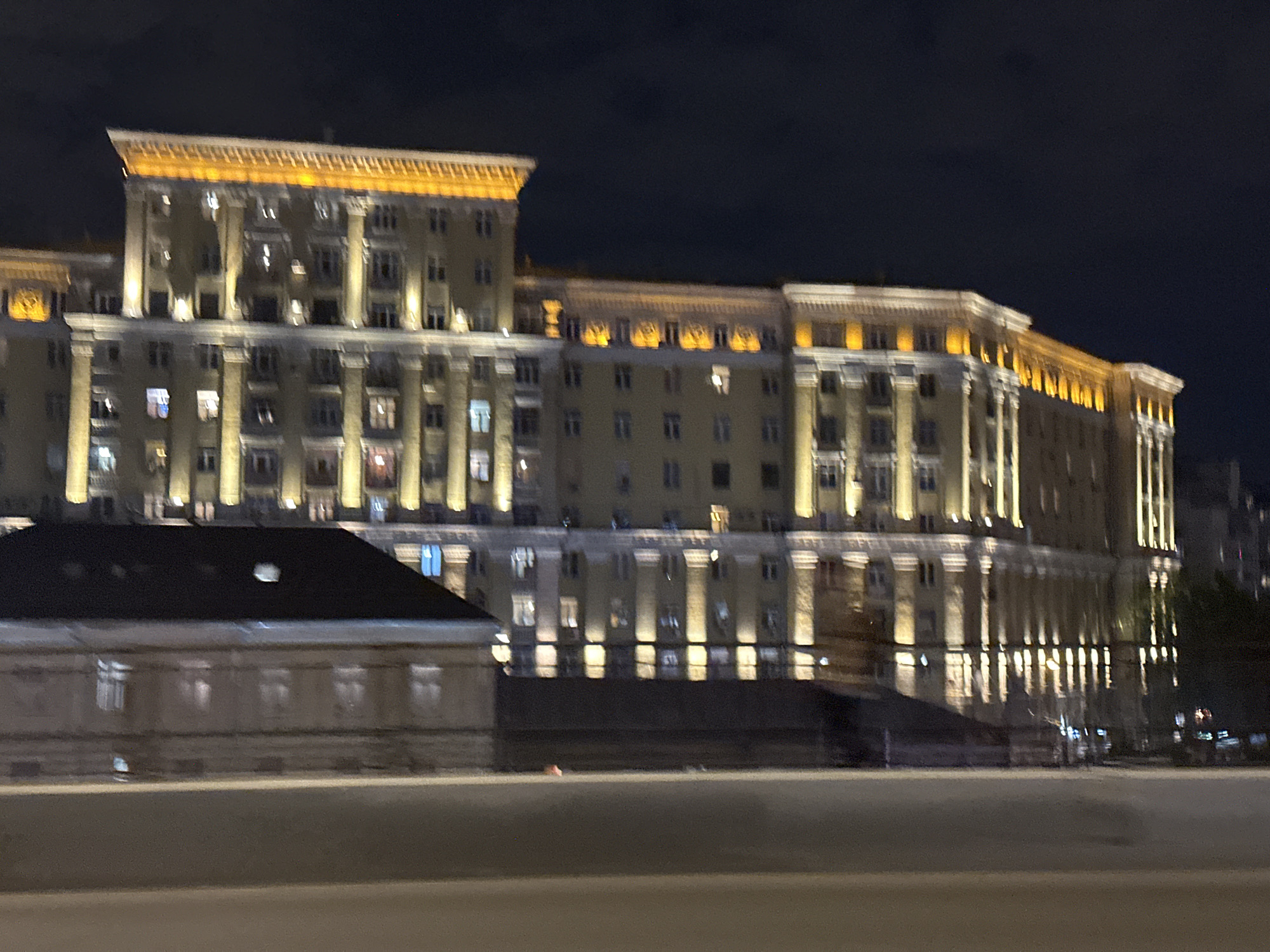 The image shows a large building illuminated at night in Moscow, Russia. The building has a Stalinist architectural style, with multiple floors, rows of windows, and decorative columns. The top of the building is brightly lit with a warm, yellow glow. In front of the main building is a smaller, darker building with a simple, sloping roof. The foreground is out of focus, showing what appears to be a road or sidewalk. The overall scene has a blurry, nighttime quality. The Stalinist style points to one of the "Seven Sisters" skyscrapers in Moscow.
