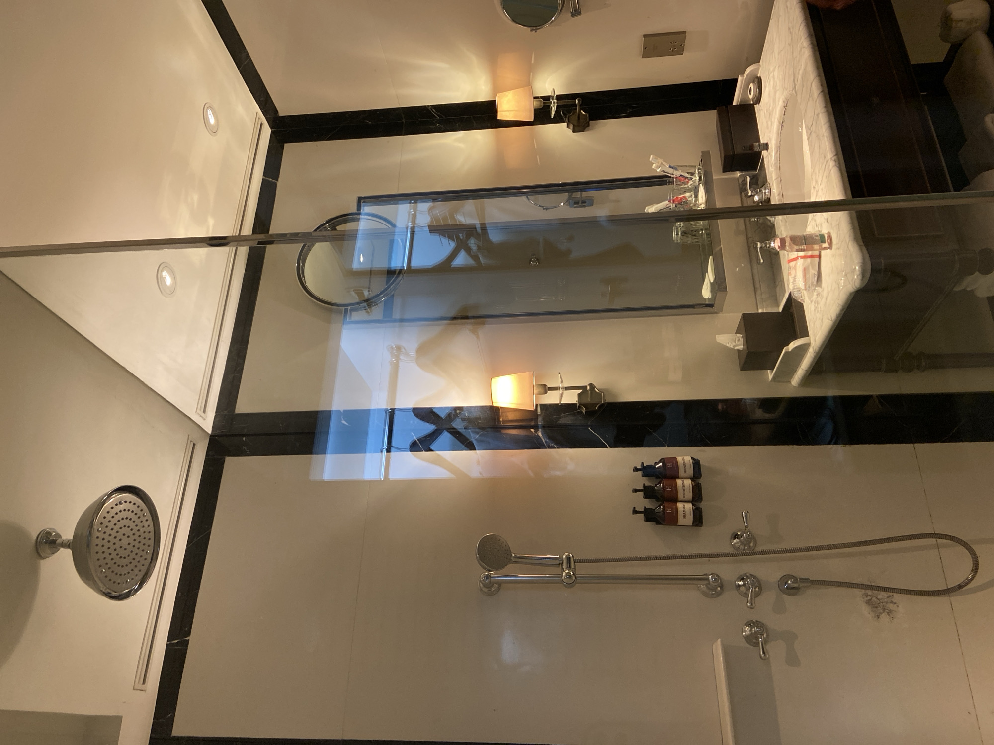 The image shows a modern bathroom with a marble countertop and sink. A large mirror is mounted above the sink, reflecting a wall sconce and toothbrushes in a holder. Several toiletries are also visible on the counter. In the foreground, there is a shower head and a handheld shower wand. The overall impression is one of a clean and well-appointed bathroom, possibly in a hotel or luxury residence. The lighting suggests it is evening or interior lighting is on. The location context is Central Jakarta, Indonesia. There is no visible text in the image.
