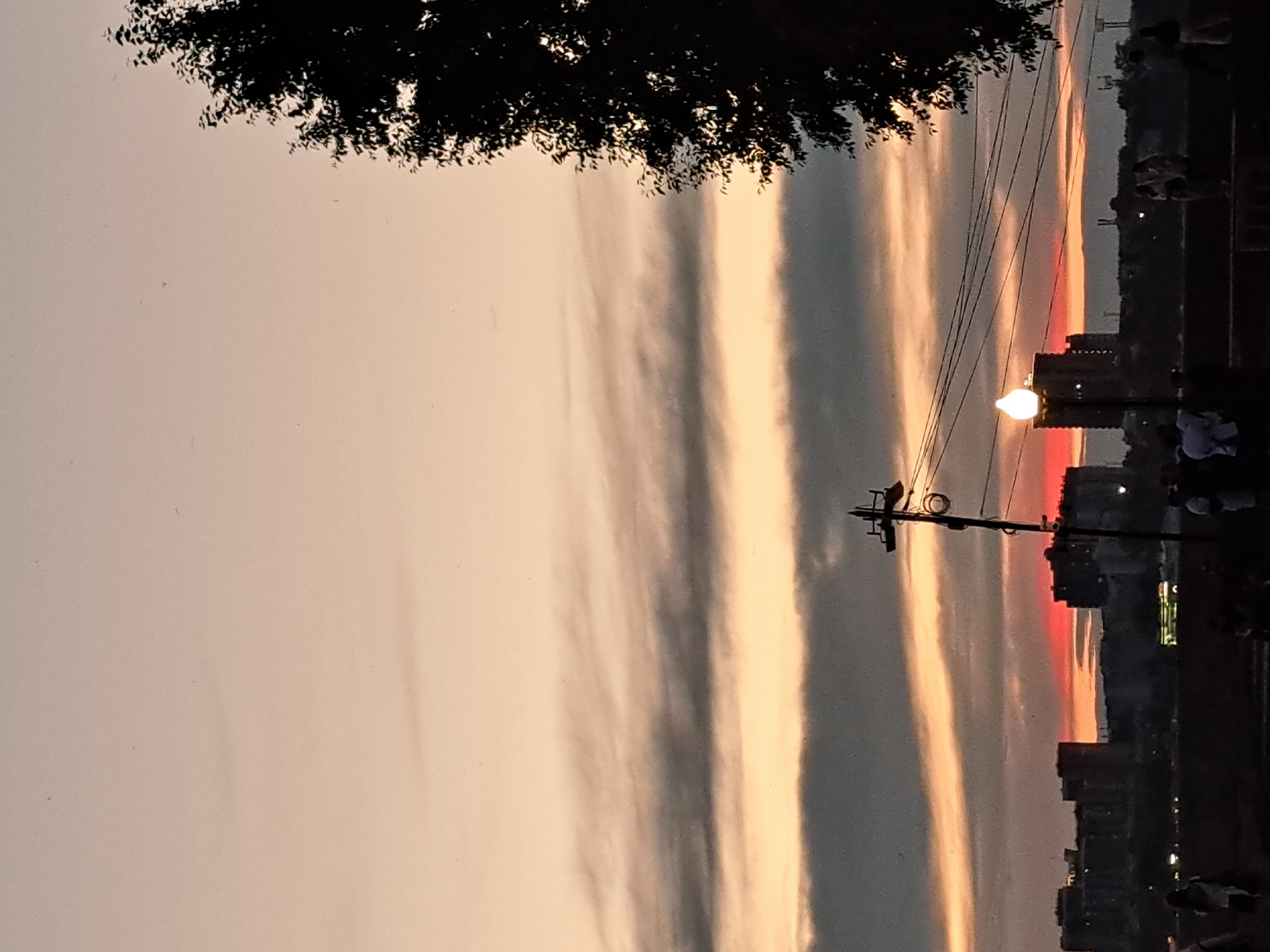 The image captures a silhouetted cityscape at sunset, likely from a vantage point overlooking the Angara River in Irkutsk, Russia. The sky is painted with hues of orange and pink, with layers of clouds creating a textured effect. In the foreground, a dark silhouette of people can be seen gathered, suggesting a public area like a promenade or embankment. To the right, a large tree with dense foliage is silhouetted against the sky. In the midground, a row of buildings forms the city's skyline, some appearing to be apartment blocks. A streetlamp with a glowing bulb is prominently featured, casting light on the scene. Utility poles and power lines are visible, crisscrossing the sky. The overall atmosphere is serene, with the fading light of day creating a warm and tranquil ambiance. The specific location within Irkutsk is not explicitly identified but the view of the river and city buildings is consistent with areas along the Angara.