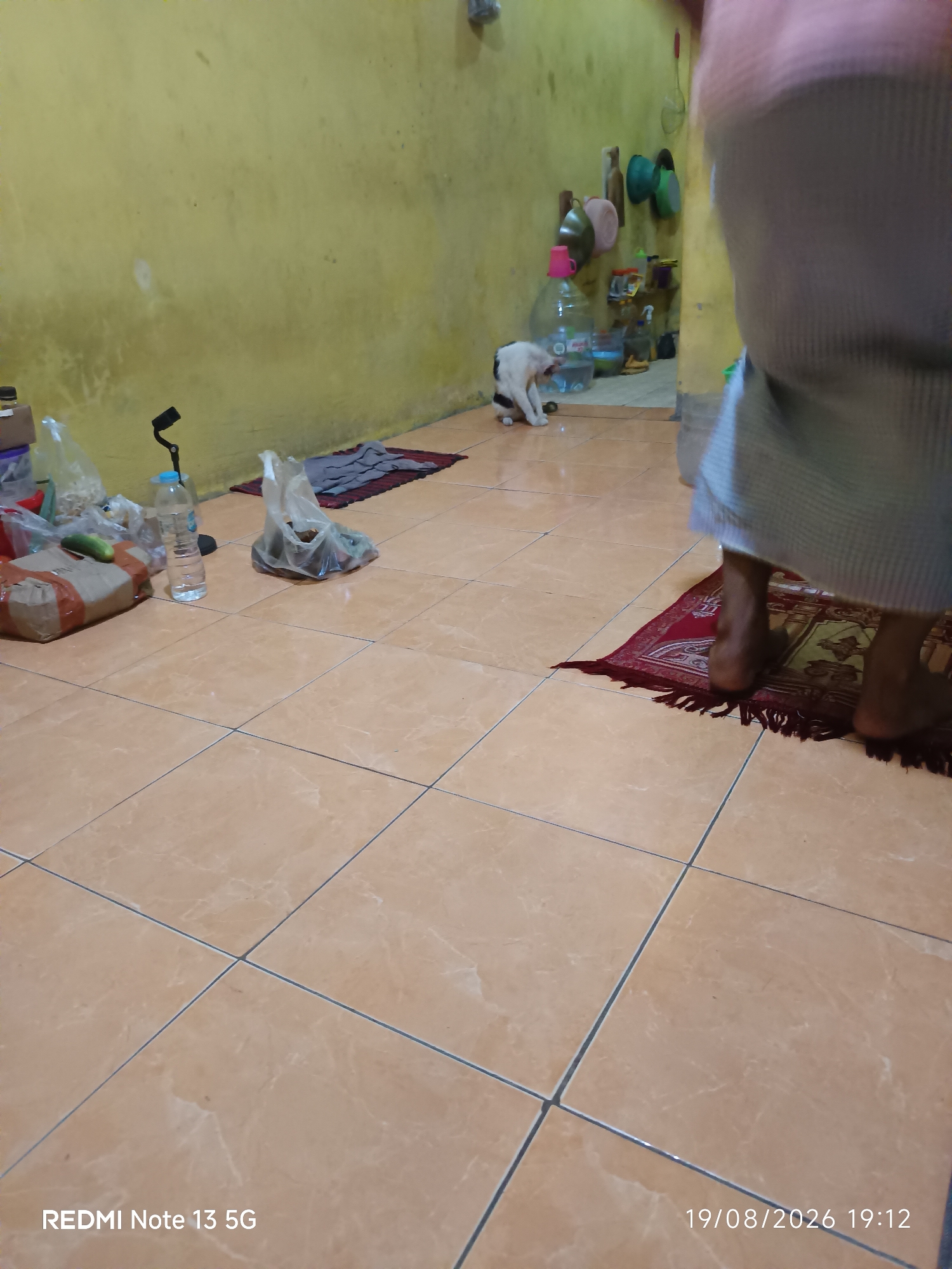 The image depicts an indoor scene, likely a dwelling, with light brown tiled flooring. On the left side, there are several items, including a brown cardboard box, a plastic bag containing what appears to be food or goods, a bottle of water, and a black object attached to a stand. In the middle ground, a white and black cat is visible, facing left. Further in the background, a yellow wall is prominent, with various items hanging on it, such as hats or baskets. There are also large plastic bottles, one with a pink cap, and shelves with containers. On the right side of the image, a person's legs and feet are visible, wearing a light-colored garment. The person is standing on a small, red rug with fringe. The timestamp "19/08/2026 19:12" and "REDMI Note 13 5G" are present in the lower right and lower left corners respectively, indicating the image was taken on August 19, 2026, at 7:12 PM. The location is specified as Talaga Sari, Indonesia.