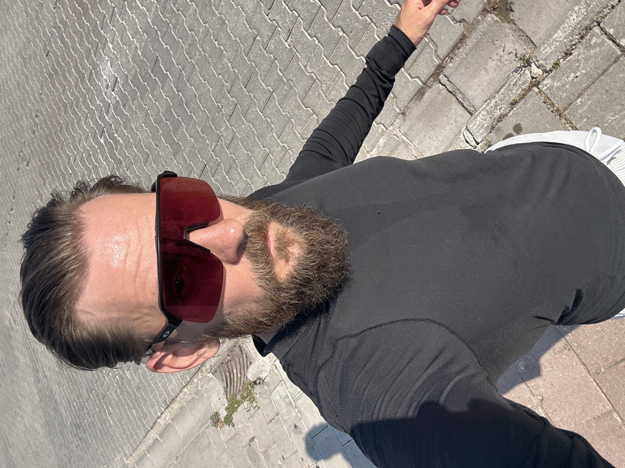 A man with a beard and sunglasses, wearing a black long-sleeved shirt and white shorts, stands on a paved walkway in Fethiye, Turkey. The walkway is made of interlocking gray pavers. Sunlight casts a shadow of the man on the walkway. The man is looking at the camera, and appears to be holding the camera out to take a selfie. A drainage grate is visible on the left side of the image, with small plants growing in the cracks between the pavers. The background is a blur of the paved walkway.