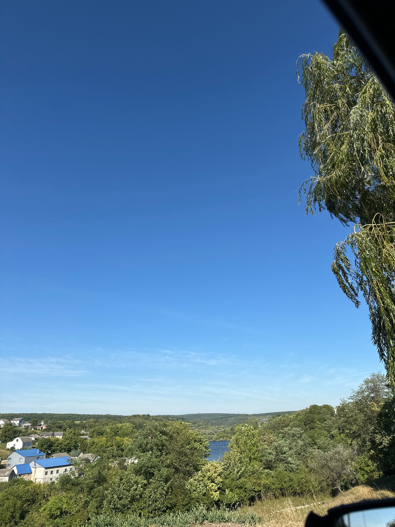 The image captures a rural landscape with a clear blue sky. In the foreground on the right, a weeping willow tree hangs down, its branches obscuring part of the sky. Below the sky, rolling green hills are dotted with trees. In the mid-ground, a small village is visible, featuring houses with blue roofs, interspersed with greenery. A body of water, likely a river or lake, can be seen in the distance. The overall scene suggests a serene, natural setting in the countryside, possibly in or near the city of Makiv, Ukraine, given the context. The image appears to be taken from a slightly elevated position, looking down onto the village and the surrounding landscape.