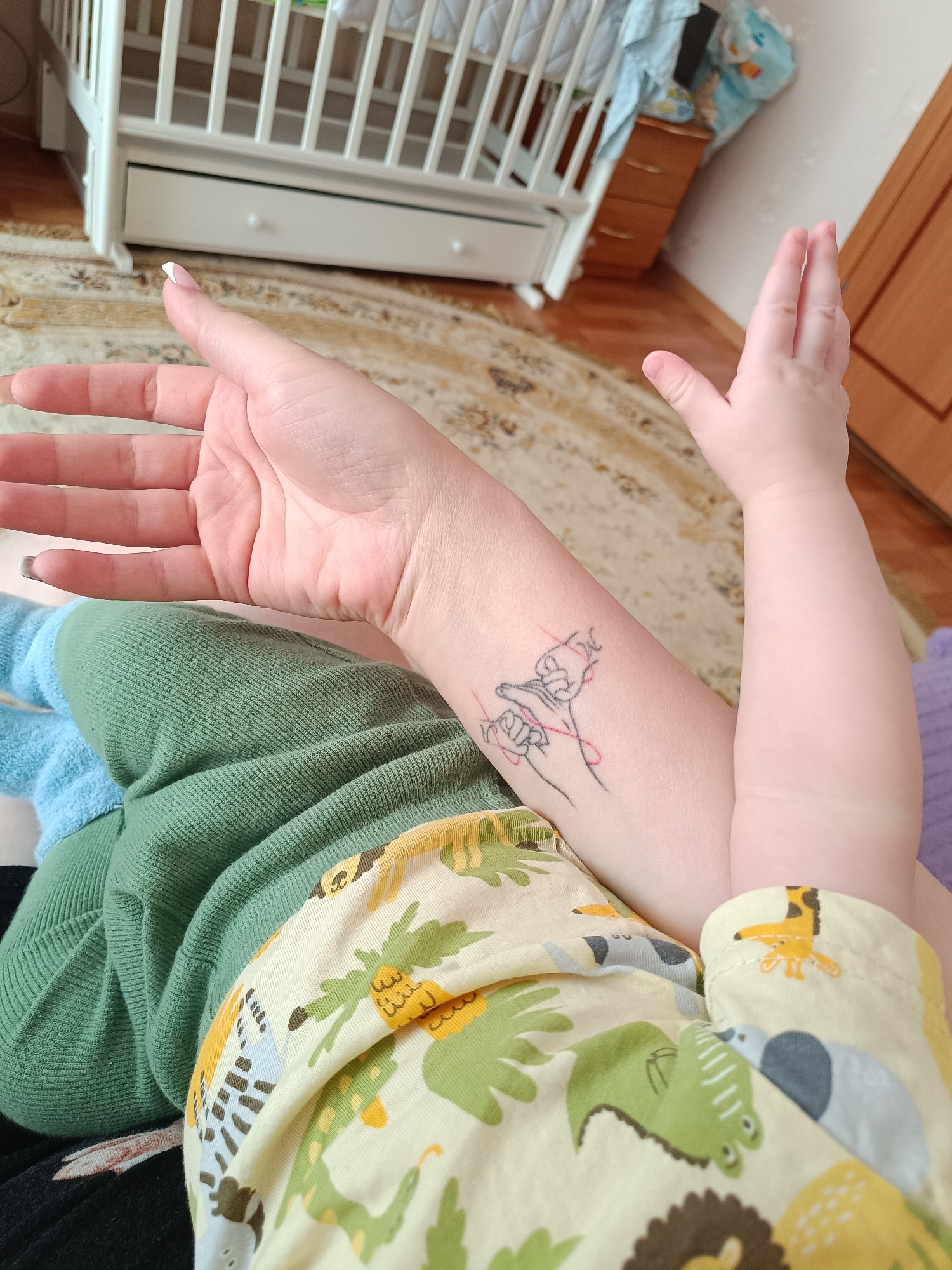 A high-angle medium shot depicts a person's arm with a tattoo on the forearm, alongside a baby's hand reaching upwards. The person's hand has a white manicure, and the arm also features a tattoo of interlocked hands connected by a red line. The baby's hand is in the upper right of the frame, reaching up. The clothing worn by the person is green and black. The baby's clothing is yellow with an animal print. A baby crib and a dresser are partially visible in the background. The image was likely taken indoors. No information about the location in Tobolsk, Russia, can be determined from the image.