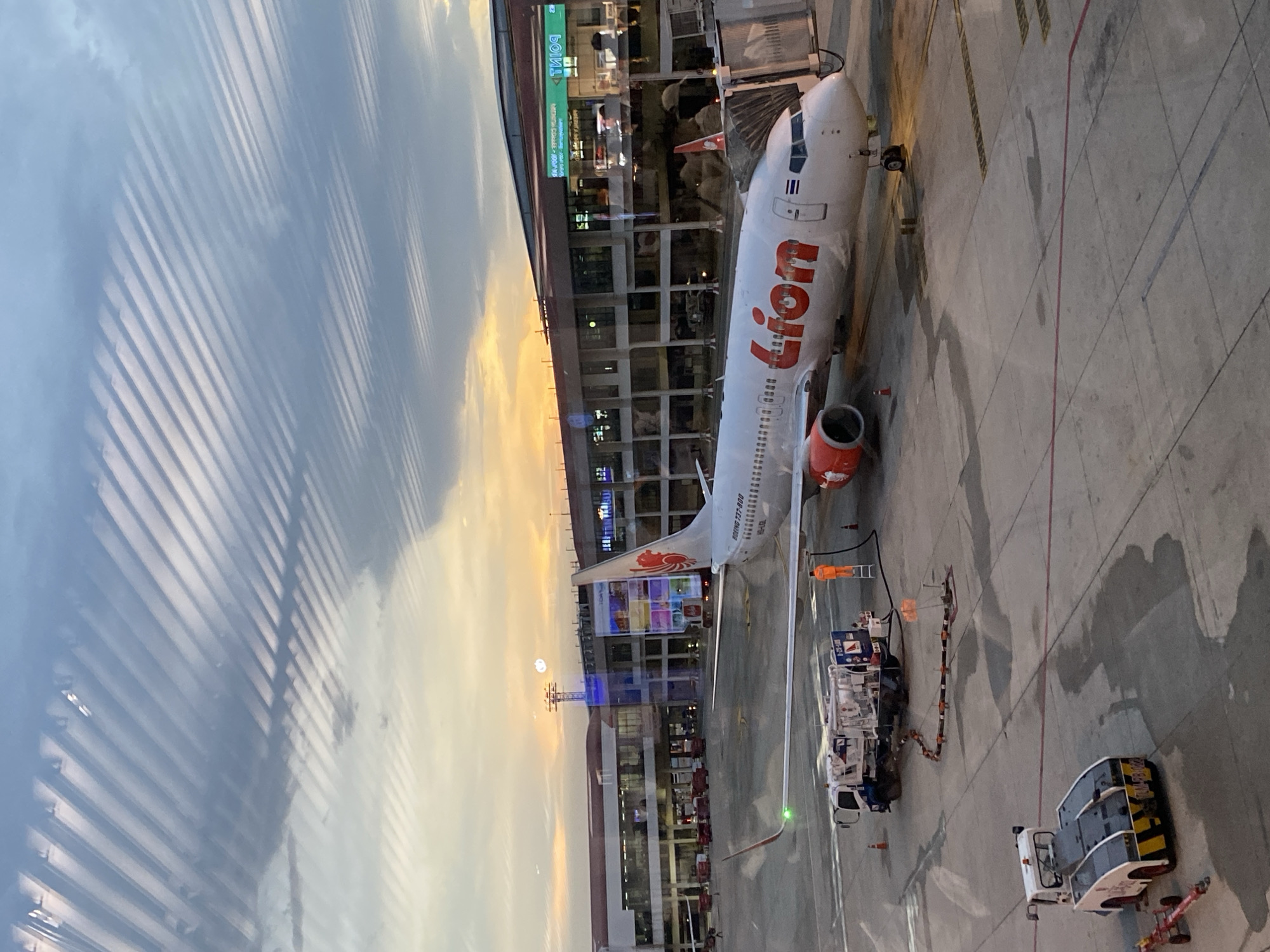 The image shows a Lion Air Boeing 737-800 aircraft parked at an airport gate, presumably Suvarnabhumi Airport in Bangkok, Thailand, given Lion Air's presence there. A fuel truck is connected to the aircraft, indicating refueling operations. A baggage handling vehicle is nearby. The scene appears to be viewed from inside the terminal, through a window with visible reflections. The sky is partially cloudy with the setting sun visible.