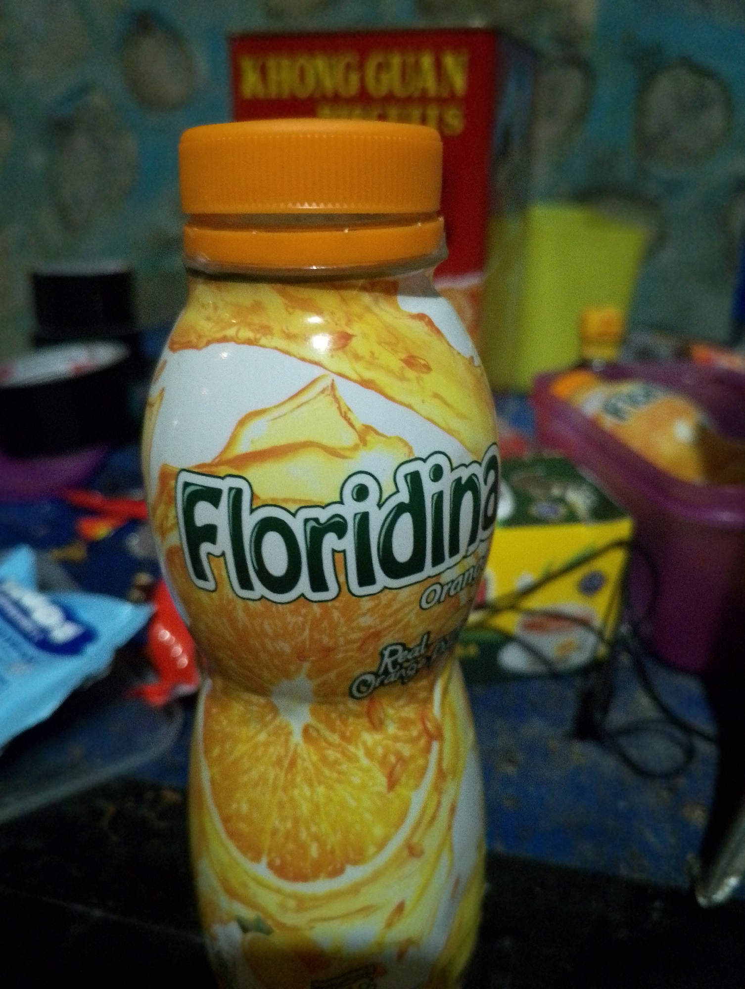 A close-up, eye-level shot shows a bottle of Floridina orange juice. The bottle has an orange cap and the label features a graphic of oranges and the brand name "Floridina" in green lettering. The background is blurry and shows various objects, including a red container with text in yellow, a yellow pitcher, and a purple container. The overall scene appears to be indoors, possibly in a home or shop. It is not possible to determine the location in Tomohon, Indonesia from the image alone.