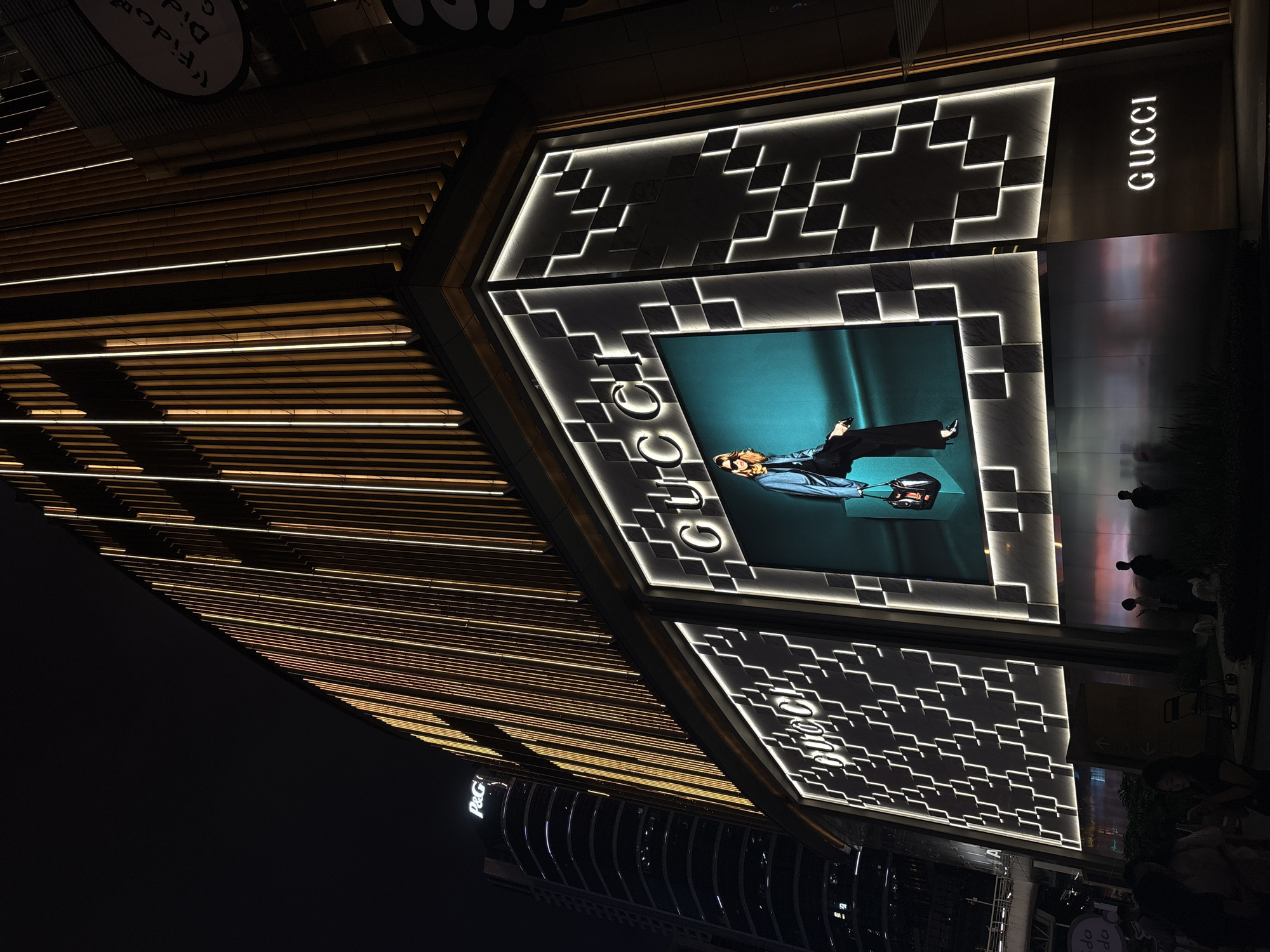 This is a nighttime, low-angle, exterior shot of a modern building facade, likely a luxury retail store, in Tianhe District, China. The building features a striking architectural design with horizontal wooden slats illuminated by warm interior lights, creating a striped effect that angles upwards. Above this, the facade is adorned with geometric patterns of white lights set against a dark, textured background. A large digital screen displays a fashion advertisement featuring a model in a dark suit and heels, holding a handbag, against a teal backdrop. The brand "GUCCI" is prominently displayed in white illuminated letters on the upper right side of the facade. In the lower right corner, several shadowy figures of people are visible, suggesting it is a public area with pedestrian traffic. The "P&G" logo is also visible in white illuminated letters on a lower section of the building. The overall impression is one of high-end commercial sophistication and a vibrant urban atmosphere at night.