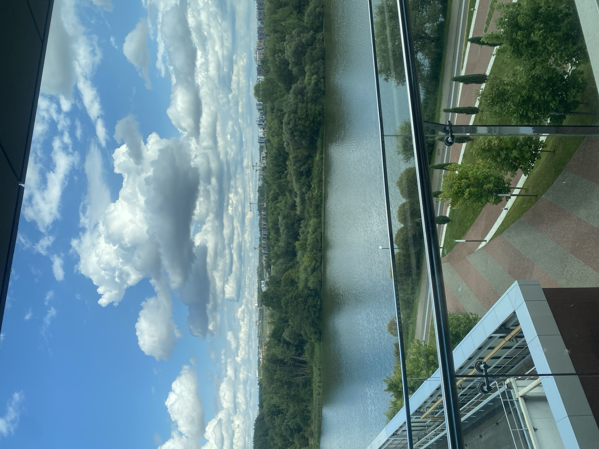 The image depicts a view from an upper floor of a building, looking out over a river, a treeline, and a cityscape under a partly cloudy blue sky. In the foreground, a glass balcony railing is visible. The river is wide and calm, reflecting the sky. The treeline is dense with various shades of green, suggesting a mix of deciduous and evergreen trees. Beyond the trees, a modern cityscape with apartment buildings and construction cranes can be seen. The foreground also shows a paved area with patterned brickwork and landscaping, including manicured grass and rows of cypress trees. This view is consistent with locations along the Svislach River in Minsk, Belarus, particularly areas undergoing development or adjacent to residential complexes. There are no people visible in the image. The overall scene is one of a developed urban area integrated with natural elements, suggesting a contemporary setting.