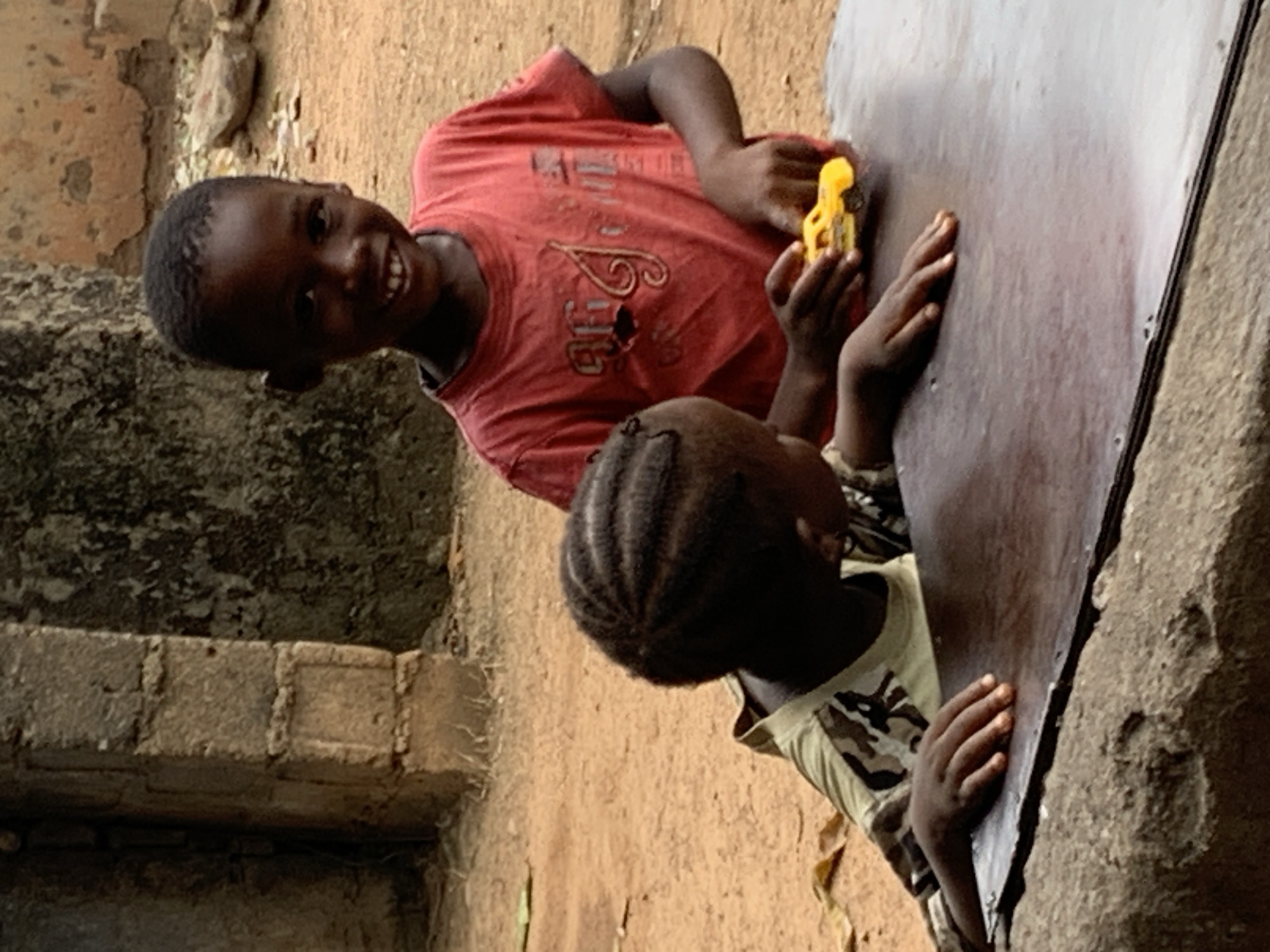 Two dark-skinned children are playing with a yellow toy car on a wooden surface in Akwanga, Nigeria. One child, wearing a red t-shirt, is looking towards the camera and smiling while holding the toy car. The other child, wearing a camouflage patterned shirt and with their hair in braids, has their hands on the wooden surface. The environment includes a mud wall, a stone structure, and a dirt ground.
