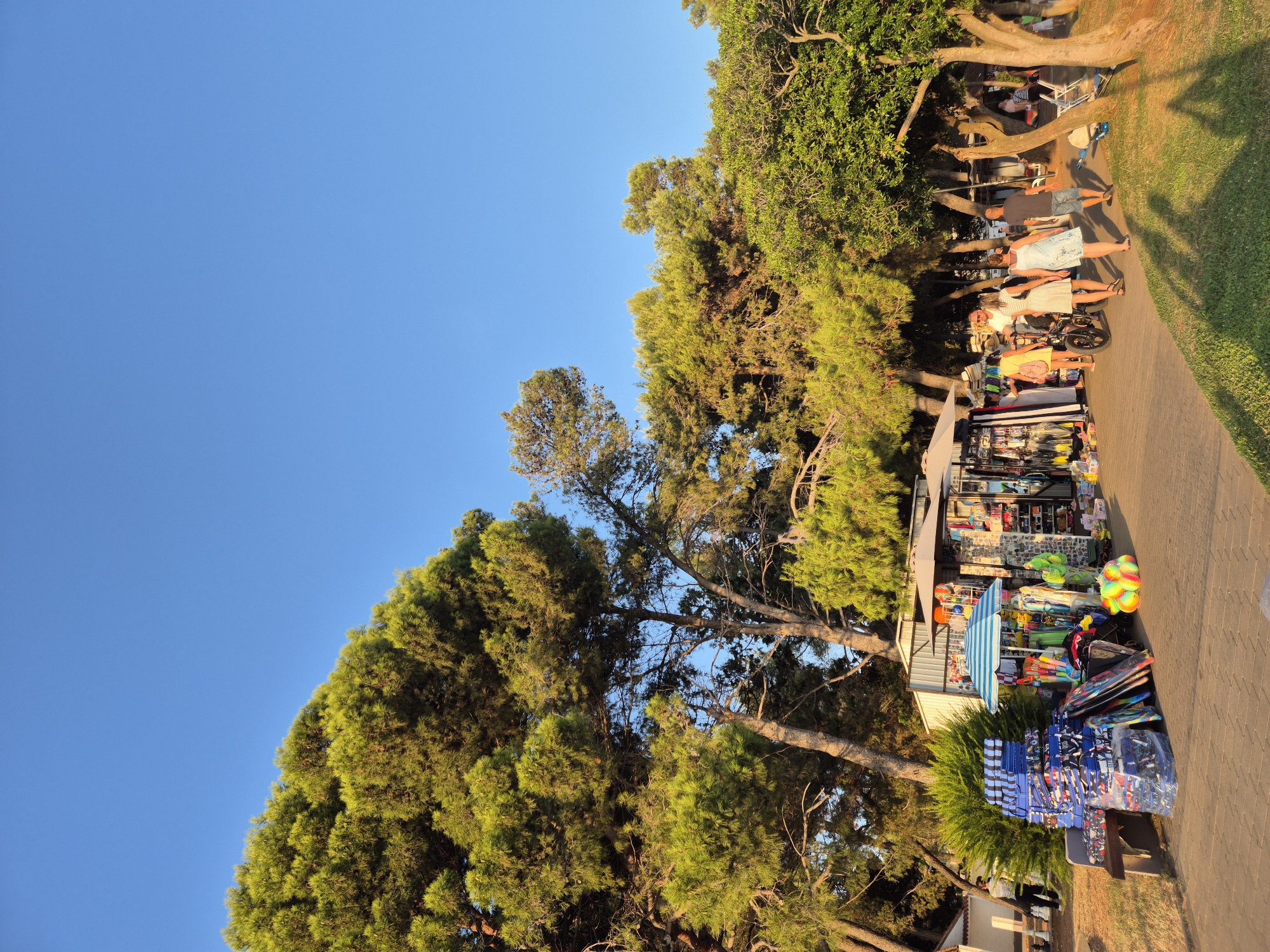 In Katoro, Croatia, a seaside path is lined with trees and a small market stall selling beach accessories. Several people are browsing the merchandise, including beach mats, umbrellas, and inflatable toys. The sun is setting, casting long shadows across the path and the surrounding grassy area. The sky is clear and blue. The market stall is colorful and inviting, with a variety of goods displayed under a striped umbrella. A few people are walking away from the stall, while others are examining the items. The overall scene is relaxed and casual, indicative of a vacation destination.