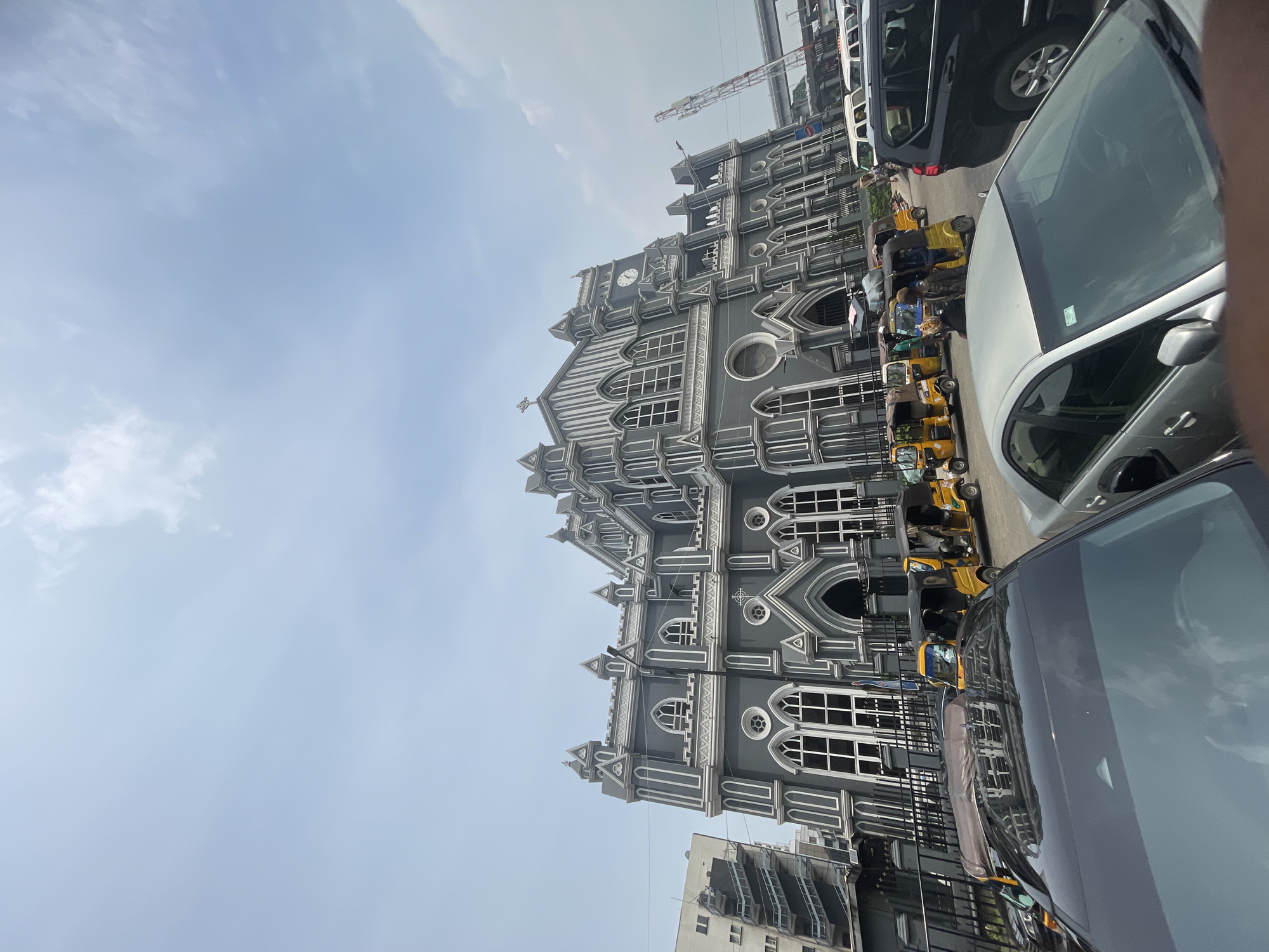 The image shows the Cathedral Church of Christ in Lagos, Nigeria. The gray building with white trim is viewed from ground level, with cars and yellow "Keke" auto-rickshaws in the foreground. The sky is light blue with some clouds. People are seen inside the auto-rickshaws, but their actions are indiscernible.