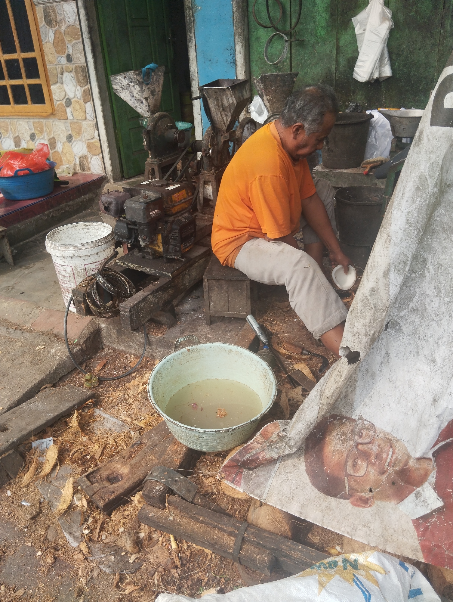 The image depicts an outdoor scene in Karangdoro, Indonesia, likely a workshop or processing area. In the foreground, a man, appearing to be of middle age, is seated on a wooden crate. He is wearing an orange t-shirt and light-colored pants. The man is holding a halved coconut in his right hand and appears to be in the process of preparing it. In front of him, a light blue bucket contains murky water, and scattered around the ground are coconut husks and debris.

Behind the man, several pieces of machinery are visible, including what appears to be a small engine connected to a grinding or milling apparatus. There are also various buckets and containers present. A large, white, torn tarp or banner is prominently positioned on the right side of the frame, partially obscuring a printed image of a man's face with glasses. The background shows a building with tiled walls and a green door, suggesting a domestic or commercial establishment. The overall environment appears weathered and utilitarian, indicative of a working area.