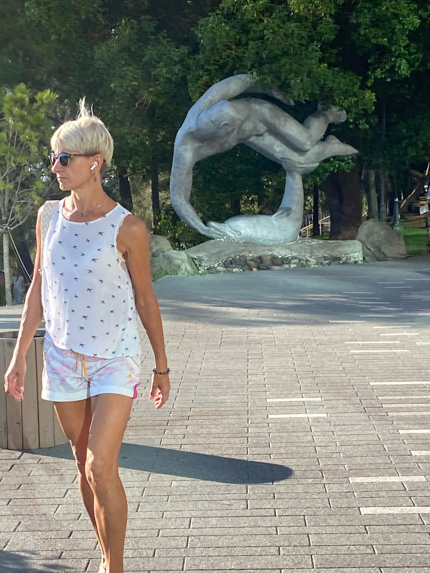 The image depicts a woman with short blonde hair and sunglasses walking on a paved pathway. She is wearing a white tank top with a bird pattern and white shorts with pink accents. The pathway is made of rectangular grey pavers, with some lighter colored strips interspersed. In the background, there is a large grey sculpture of a human figure interacting with a dolphin-like creature. Behind the sculpture are lush green trees and foliage, suggesting an outdoor park or public space. The sunlight casts a shadow of the woman on the pathway to her right. The scene appears to be a sunny day in an urban outdoor setting. The location is the central promenade of the resort city of Sochi, Russia, specifically near the dolphin monument.