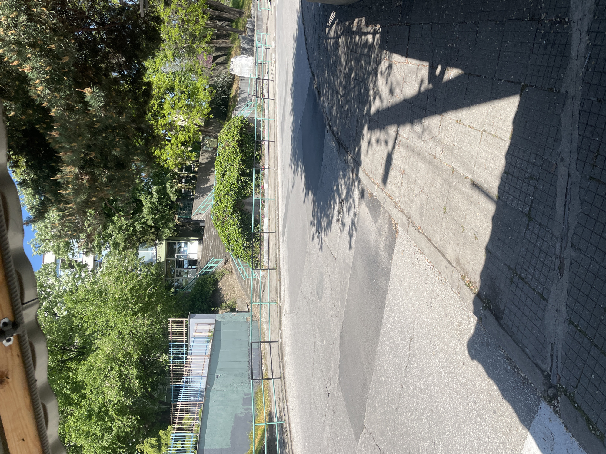 The image depicts an outdoor scene in what appears to be a residential or recreational area of Asenovgrad, Bulgaria. A paved street or pathway is visible, bordered by a raised section of land with greenery and some buildings in the background. A metallic fence with teal-colored rails runs along the edge of the raised area, suggesting a boundary between the street and the elevated terrain. The pavement consists of various types of surfaces, including asphalt and concrete tiles. Tree shadows are cast onto the ground, indicating daylight.