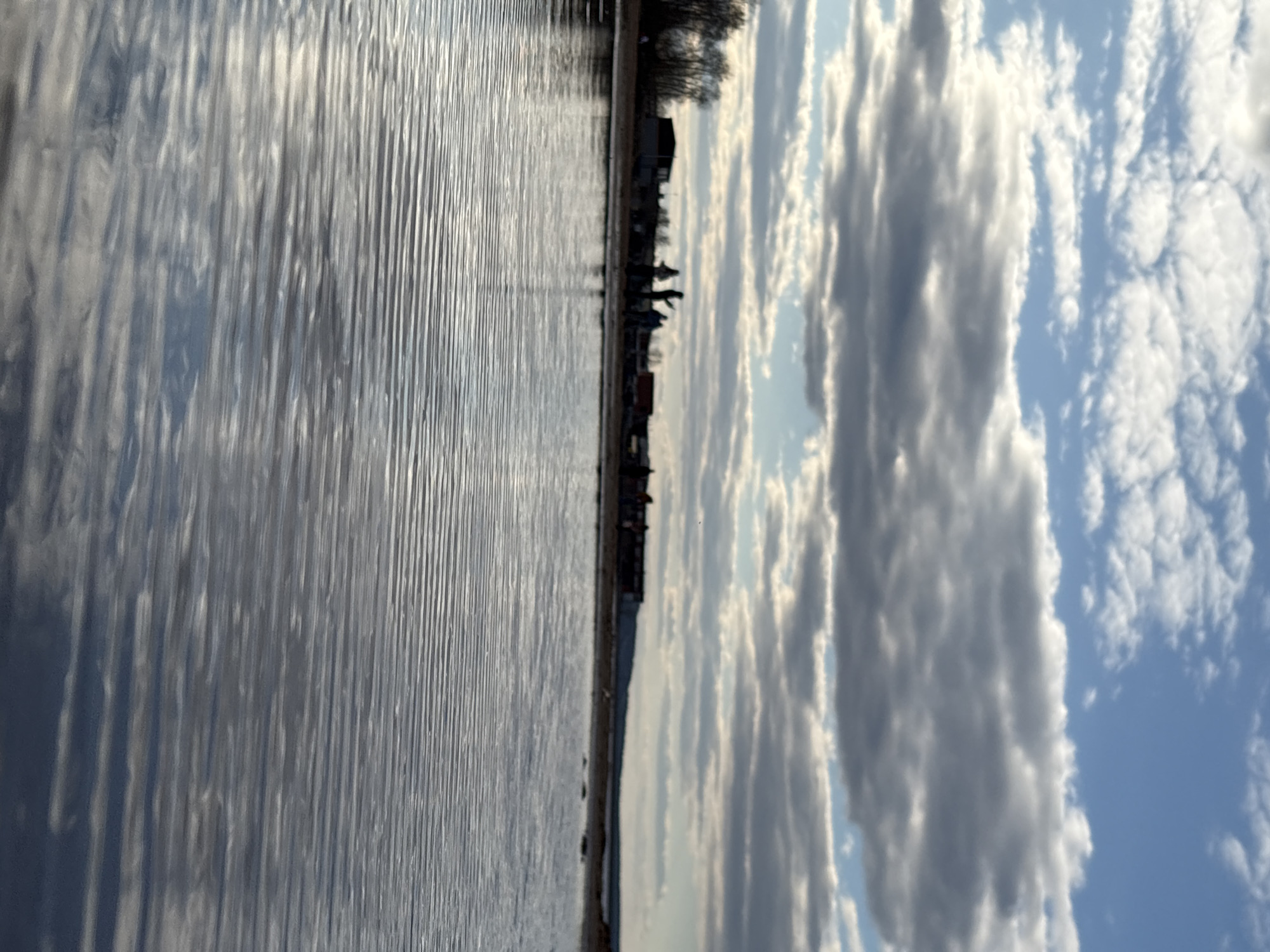 The image depicts a waterfront scene in Chaikovsky, Russia. The left side of the frame is dominated by rippled water reflecting light. The shoreline runs vertically in the center, featuring indistinct buildings and silhouetted figures of people. The right side shows a sky with scattered cumulus clouds against a blue background.