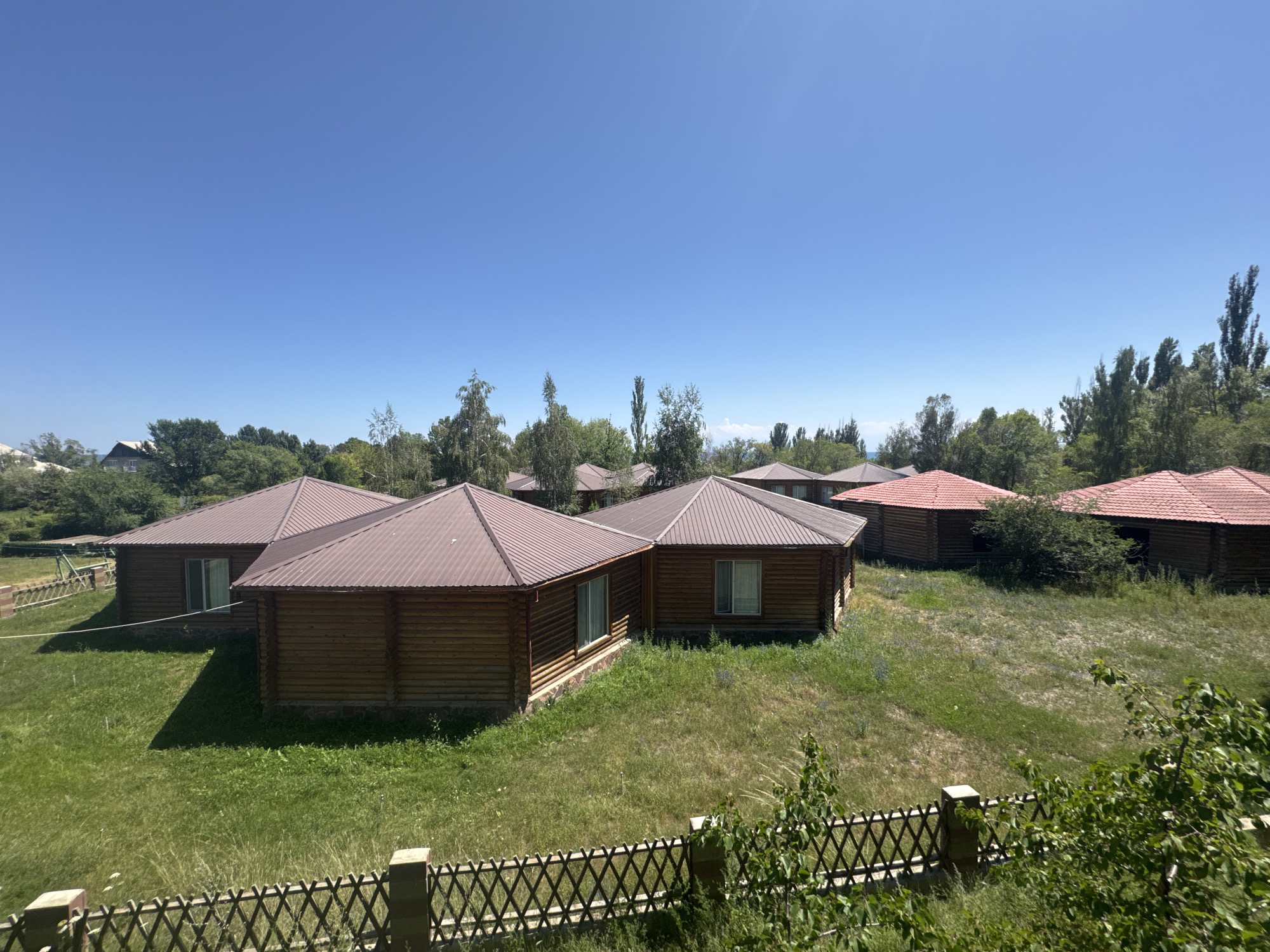 The image shows a clear blue sky over a cluster of wooden cabins with red roofs. The cabins are of a similar design, featuring log construction and large windows. They are situated on a grassy, slightly unkempt lawn, with scattered trees and bushes in the background. A wooden fence runs along the foreground of the image. The overall impression is of a rural or semi-rural setting, possibly a resort or guesthouse. The location is identified as Koshkol', Kyrgyzstan. There are no people visible in the image.