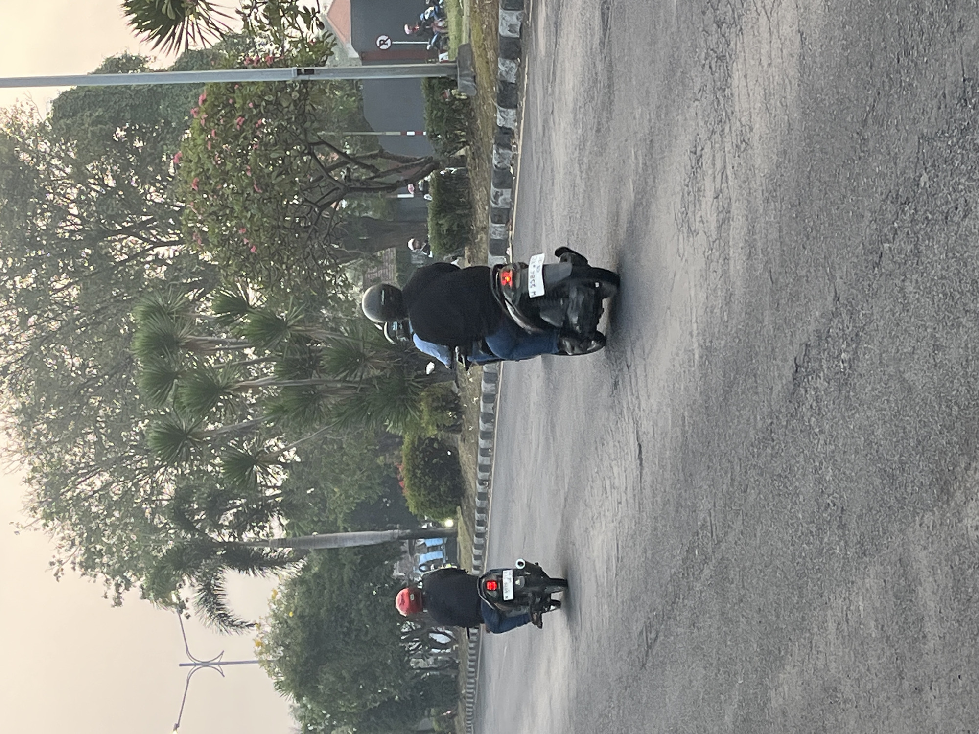 The image shows a street scene in Kludan, Indonesia. Two individuals are riding motorcycles in the foreground, both facing away from the camera. The person on the left is wearing a red helmet and a dark jacket, and the motorcycle appears to be black with a red taillight. The person on the right is wearing a black helmet and a dark jacket, riding a black motorcycle. The street is asphalt with a decorative border of black and white stones. In the background, there are various trees, including palm trees, and some buildings. The sky is overcast, suggesting it might be early morning or late afternoon. The overall environment appears to be a semi-urban or suburban area with significant greenery.