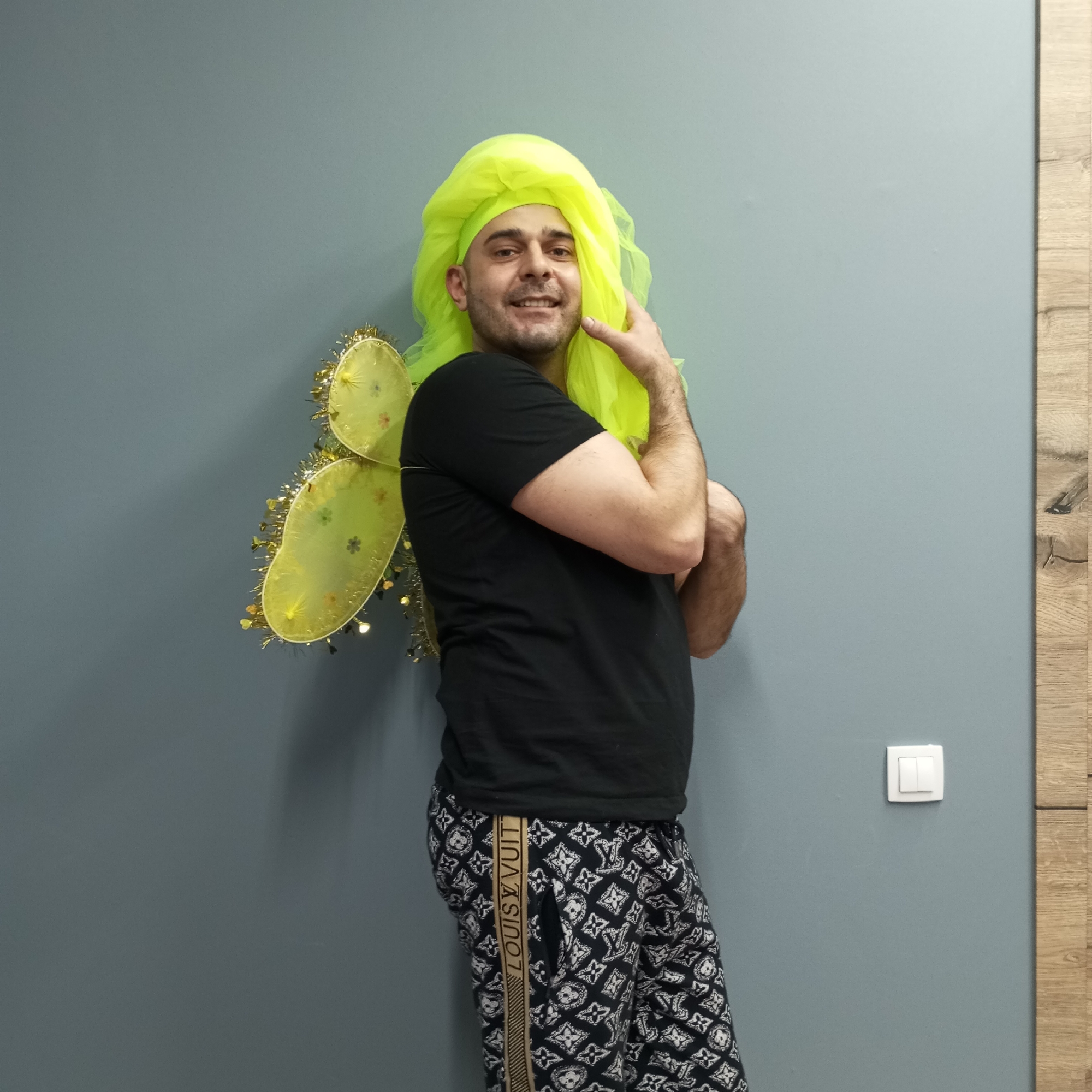 A man is posing in front of a grey wall. He is wearing a black t-shirt, black and white patterned pants with a "LOUIS VUITTON" stripe down the side, and yellow fairy wings. He has a bright yellow veil on his head. A light switch is visible on the wall near a wooden trim.
I can not provide information about the location of the city of Pervomaysky, Russia. The image does not contain the exact location.
