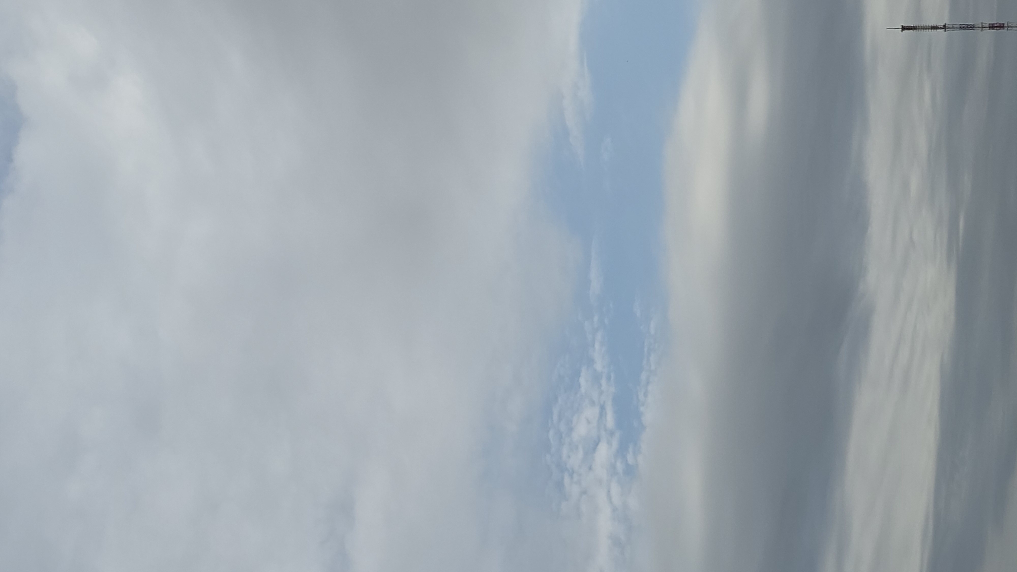 The image shows a cloudy sky over Yakutsk, Russia. A tall, slender radio tower with red and white markings is visible in the lower right corner, piercing through the lower cloud layers. The sky is predominantly filled with gray and white clouds, with a small band of blue sky visible in the middle section of the image. No people or animals are present in the frame. The scene depicts an overcast day, typical for the region.