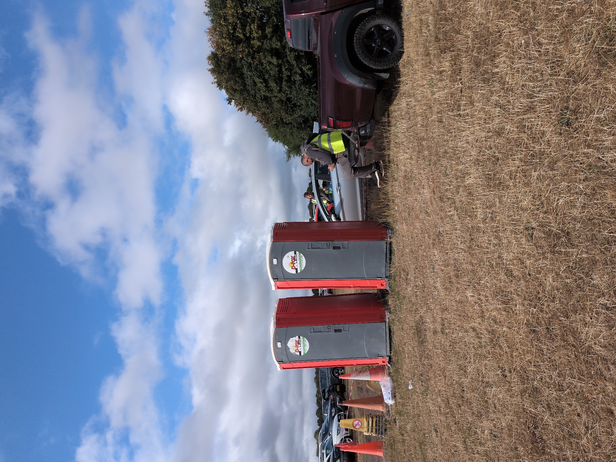 A man wearing a high-visibility yellow vest is sitting on the tailgate of a dark red pickup truck. He is looking down at his phone. Another man, also wearing a high-visibility vest, is partially visible in the driver's seat of a silver car parked next to the truck. In the foreground, two portable toilets with red frames and grey doors are set up on dry, yellowed grass. Orange traffic cones are positioned near the toilets. In the background, numerous cars are parked in what appears to be a field or large open space. The sky is blue with scattered white clouds. The location is South Staffordshire, United Kingdom.