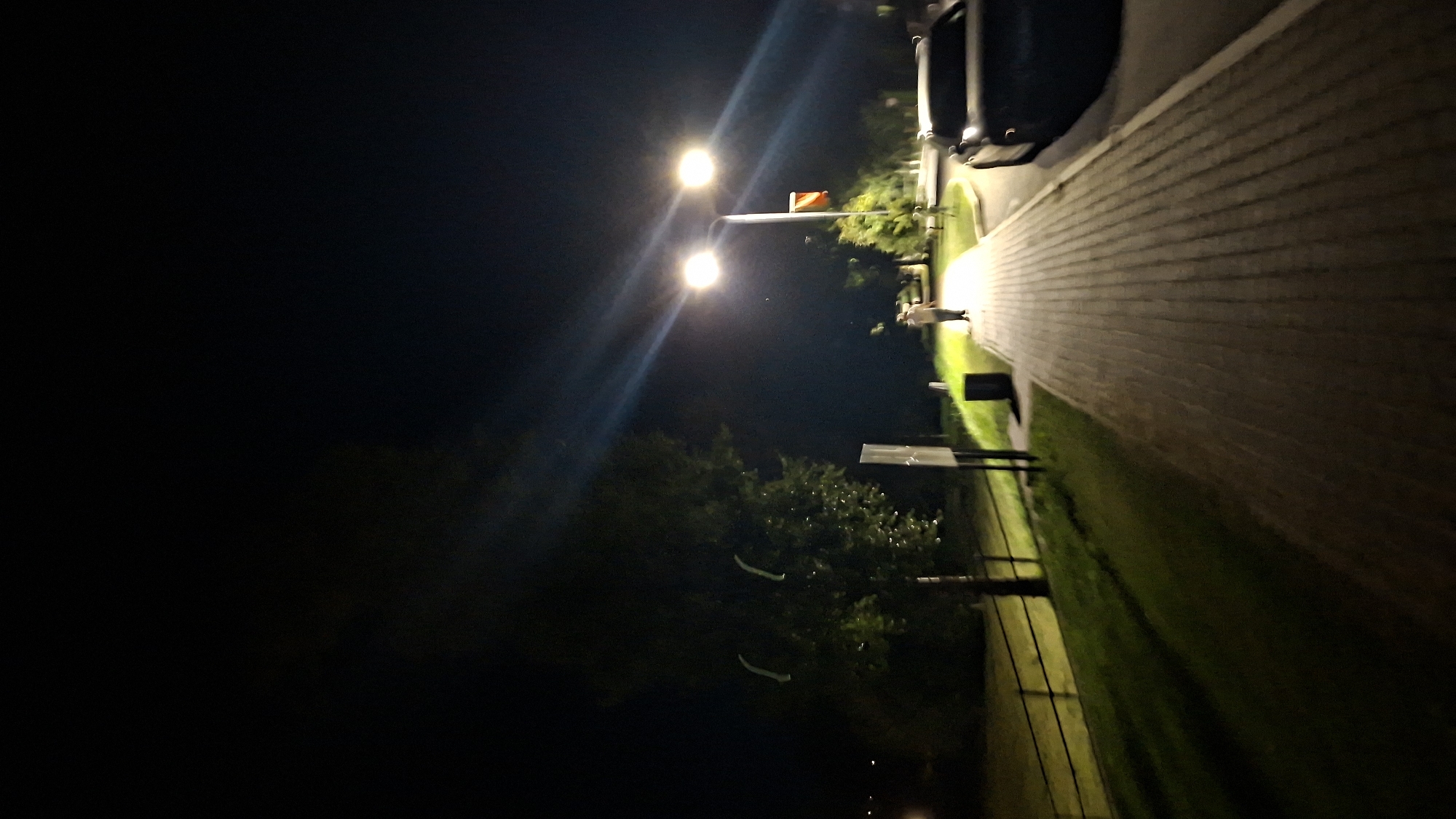 This is a night-time photograph taken on a paved pedestrian walkway, likely in the city of Myadzyel, Belarus. A tall lamppost with two bright lights illuminates the scene, casting long shadows. To the right of the walkway, a dark-colored car is parked. A lone individual, appearing to be walking away from the camera, is visible on the walkway, illuminated by the lamppost. To the left of the walkway, there is a grassy area with a black metal fence bordering it. A mature tree with green foliage is prominent in the left mid-ground. A red and white flag is attached to the lamppost. The overall environment suggests an outdoor public space at night, possibly a park or a street with pedestrian access.