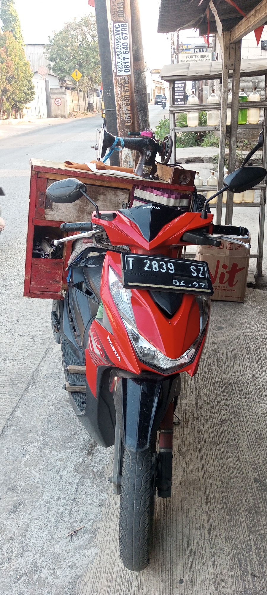 This is a front-facing, eye-level, medium shot of a red and black Honda Vario motorcycle parked on a concrete sidewalk in Setu, Indonesia. The motorcycle has a wooden box mounted behind the seat, containing a sewing machine and fabric. A black license plate reading "IT 2839 SZ" with the date "04.27" below is attached to the front of the motorcycle. To the right of the motorcycle, there is a metal shelving unit with several glass bottles containing liquids, and a large cardboard box with the "Vit" logo. Behind the motorcycle, a utility pole with various advertisements and stickers is visible. The background shows a street with trees, buildings, and a few vehicles, suggesting an urban or semi-urban environment. The lighting indicates it is daytime, possibly late afternoon due to the angle of the sun.