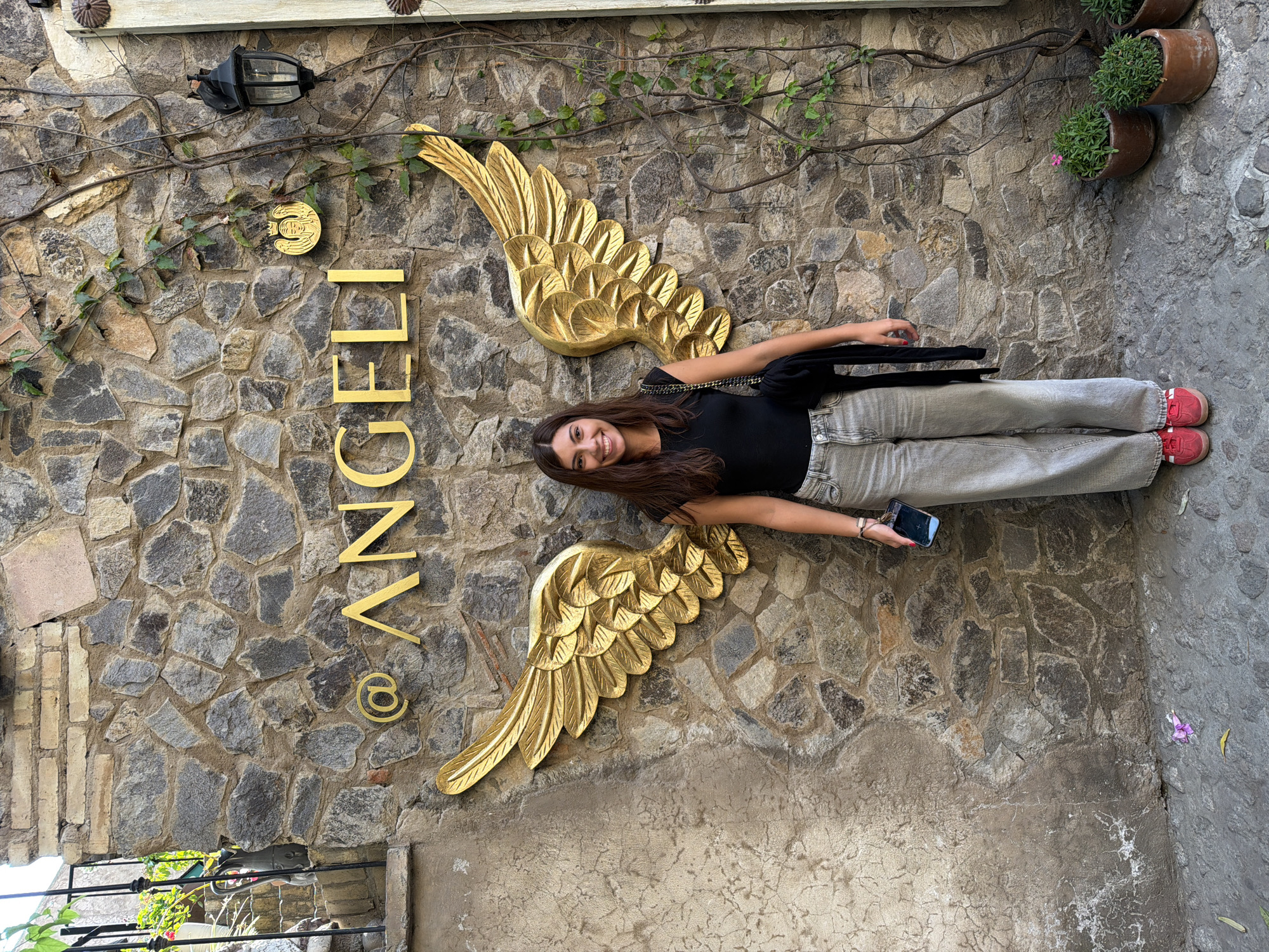 A young woman is posing in front of a stone wall that features the sign "@ ANGELI" with large golden angel wings attached to it. The woman has long brown hair and is wearing a black top and light-colored jeans, with red shoes. She holds a mobile phone in her hand. The wall is made of irregularly shaped stones, and some plants are growing on it. The location is Domingo Arenas, Mexico, based on the image context.