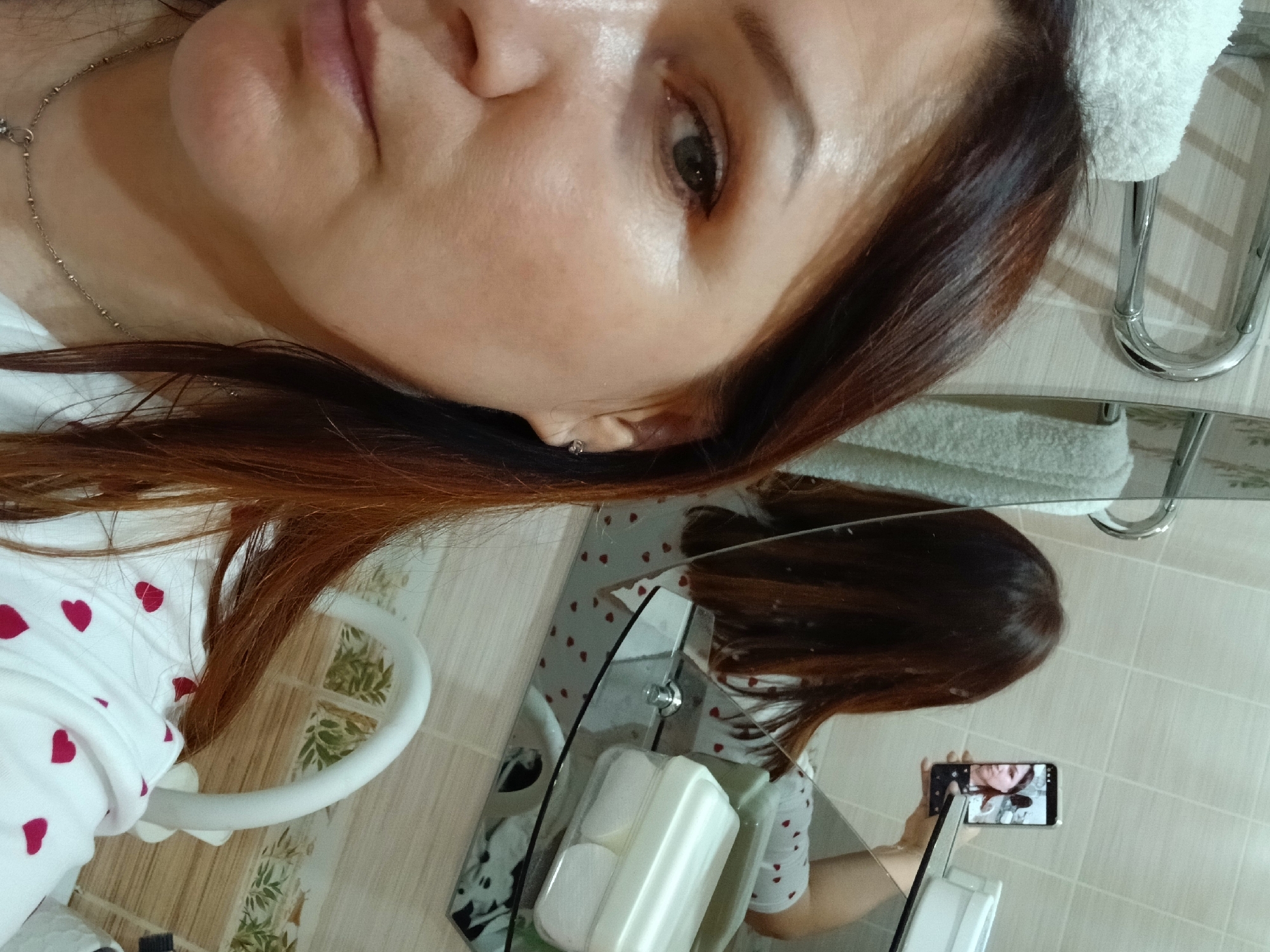 A medium-shot, indoor, close-up photograph shows a woman with medium-length, auburn hair from the shoulders up, looking into a mirror. Her left eye and cheek are the primary focus, with her hair partially obscuring her face on the right side of the frame. She is wearing a white shirt adorned with small, red hearts. In the mirror's reflection, she is holding a smartphone to take the picture, and the phone screen shows her face and upper body. The background of the image is a bathroom, with tiled walls, a chrome towel rack with towels, and a shelf holding cotton pads in a white container.
