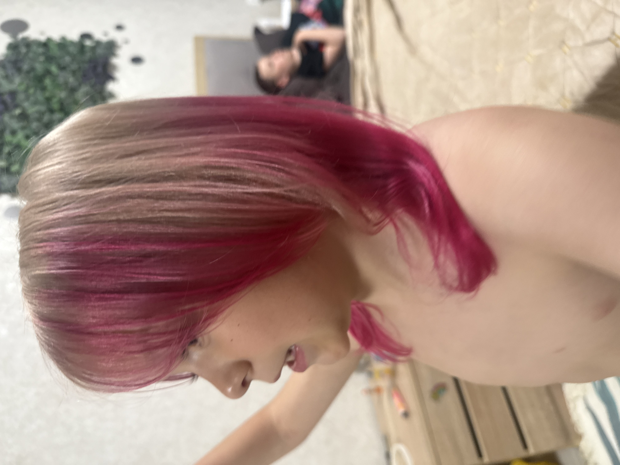 The image is a close-up shot of a fair-skinned individual, likely a child, with pink highlights in their blonde hair. The person is positioned facing slightly downward and to the right. Their mouth is slightly open. The background includes a glimpse of another person lying on a couch or bed in the upper part of the image, as well as various objects and furniture like a cabinet with decorations. A plant decoration is visible in the upper left corner of the image. The environment appears to be an interior space, possibly a home. The location within the city is not specified.
