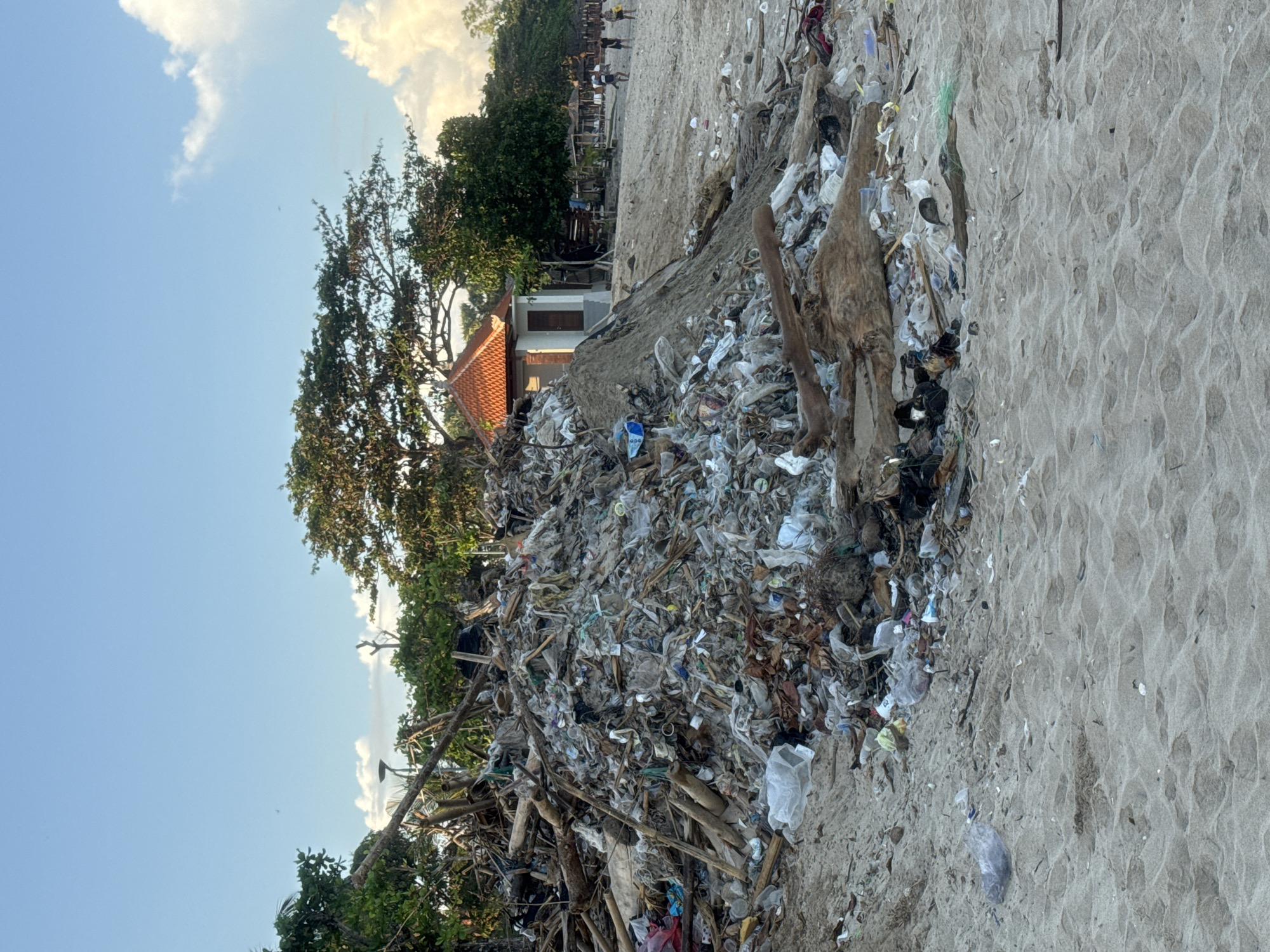 The image depicts a heavily polluted beach in Jimbaran, Indonesia. A large pile of garbage, consisting of plastic waste, wood, and debris, dominates the scene. The trash accumulation covers a significant portion of the sandy beach. To the left of the trash pile are trees and a building with a tiled roof. In the background are more trees and people on the beach. The sky is light blue with scattered clouds. The beach sand is visible in the foreground.