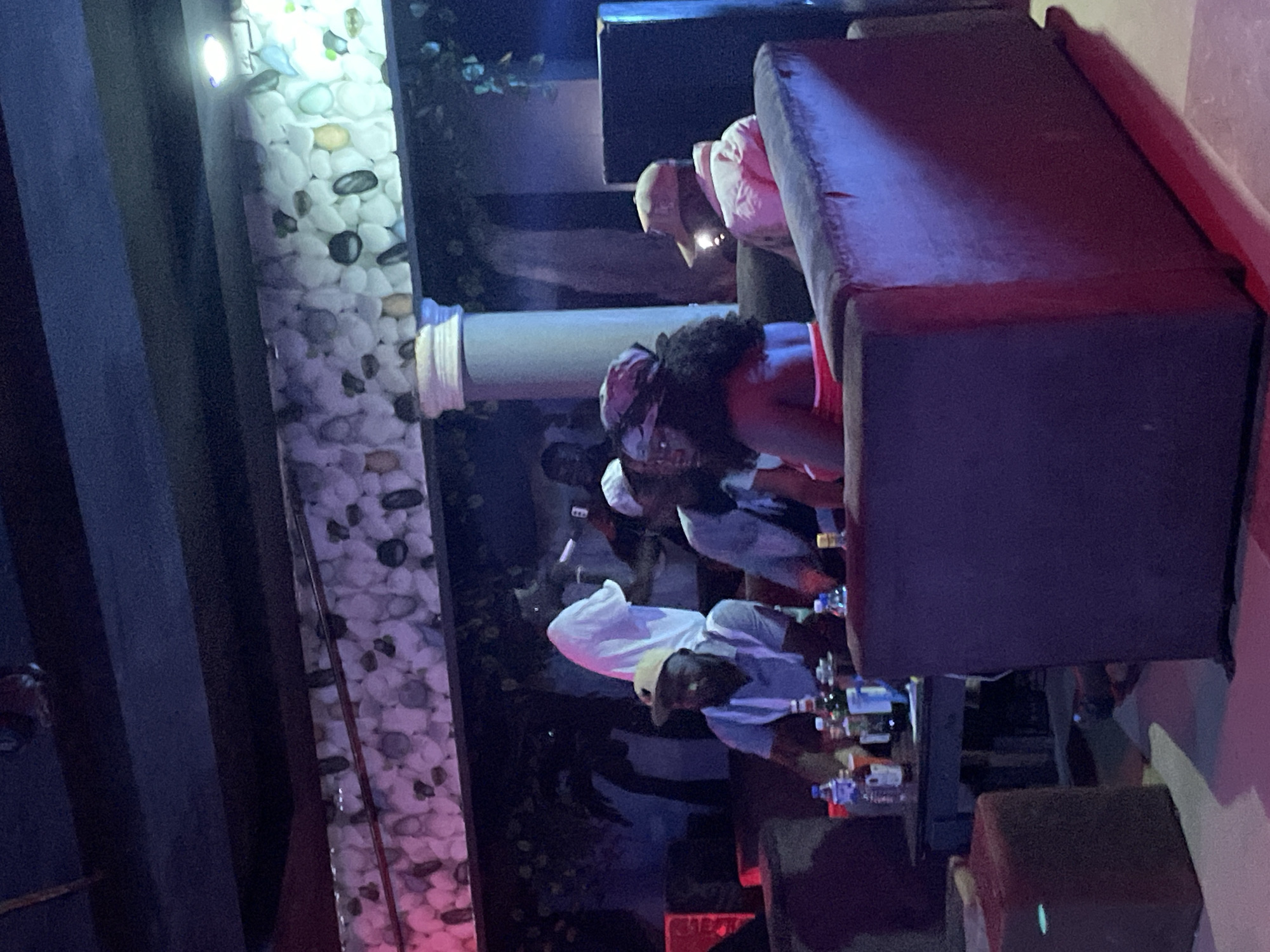 The image shows a dimly lit interior of what appears to be a lounge or club in Durumi, Nigeria. Several individuals are seated on plush, dark-colored sofas. In the foreground, a woman with dark, curly hair is wearing a red top and a headscarf. To her right, a man in a light-colored cap and jacket is looking away. To the left of the woman, a man wearing a light-colored cap and a light blue shirt is seated at a table with bottles of beverages. Behind him, another person in a white garment is visible. The background features a wall decorated with an arrangement of smooth, rounded stones in various shades of white, grey, and some with hints of color, illuminated by pink and purple lighting. A large, white pillar is also present. The overall atmosphere is relaxed, with people socializing and enjoying themselves in the indoor establishment.