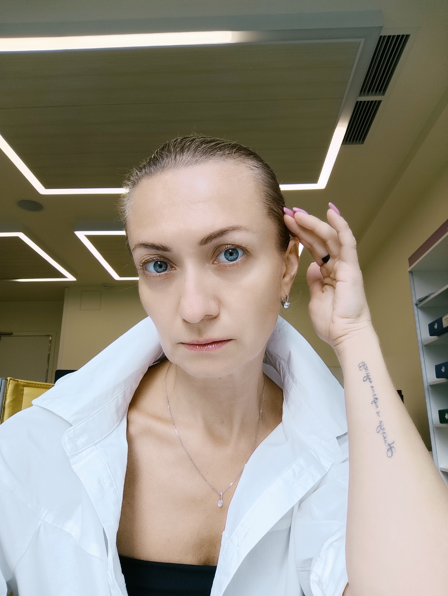 A middle-aged Caucasian woman with blonde hair pulled back and blue eyes poses for a selfie in an office environment. She is wearing a white button-down shirt and a delicate necklace with a small pendant. Her right hand is raised to her head, and a tattoo is visible on her forearm. The background features a modern ceiling with illuminated square panels and ventilation grates, as well as shelving units with files and folders. The lighting is bright, casting minimal shadows. There are no other people or clear indicators of the specific location within Moscow, Russia.