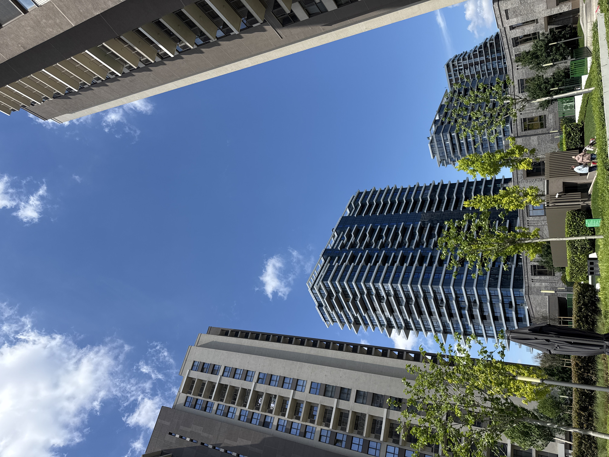 A low-angle shot shows three buildings and a clear blue sky with scattered white clouds. The building at the top-left is gray, rectangular, and features multiple balconies. The building on the bottom-left is also gray, rectangular, and has numerous windows. The building on the right is taller with a unique, stepped façade. Green trees partially obscure the bases of the buildings on the right. A grassy area with some landscaping is also visible on the right. Two people can be seen walking on the sidewalk in the background. The location is Belgrade, Serbia.