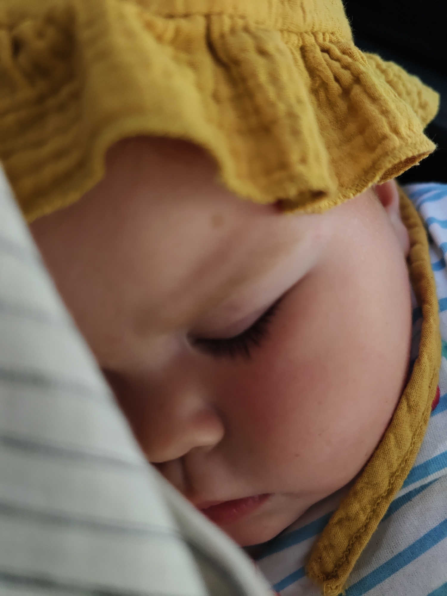 A close-up photograph shows a sleeping infant. The infant is lying on its side, with its face toward the viewer. The infant wears a yellow bonnet-style hat with a ruffled edge. The skin is light pink. Eyelashes are long and dark. The background shows striped fabric. The location is not identifiable from the image, and there is no information that connects the image to Chaikovsky, Russia.