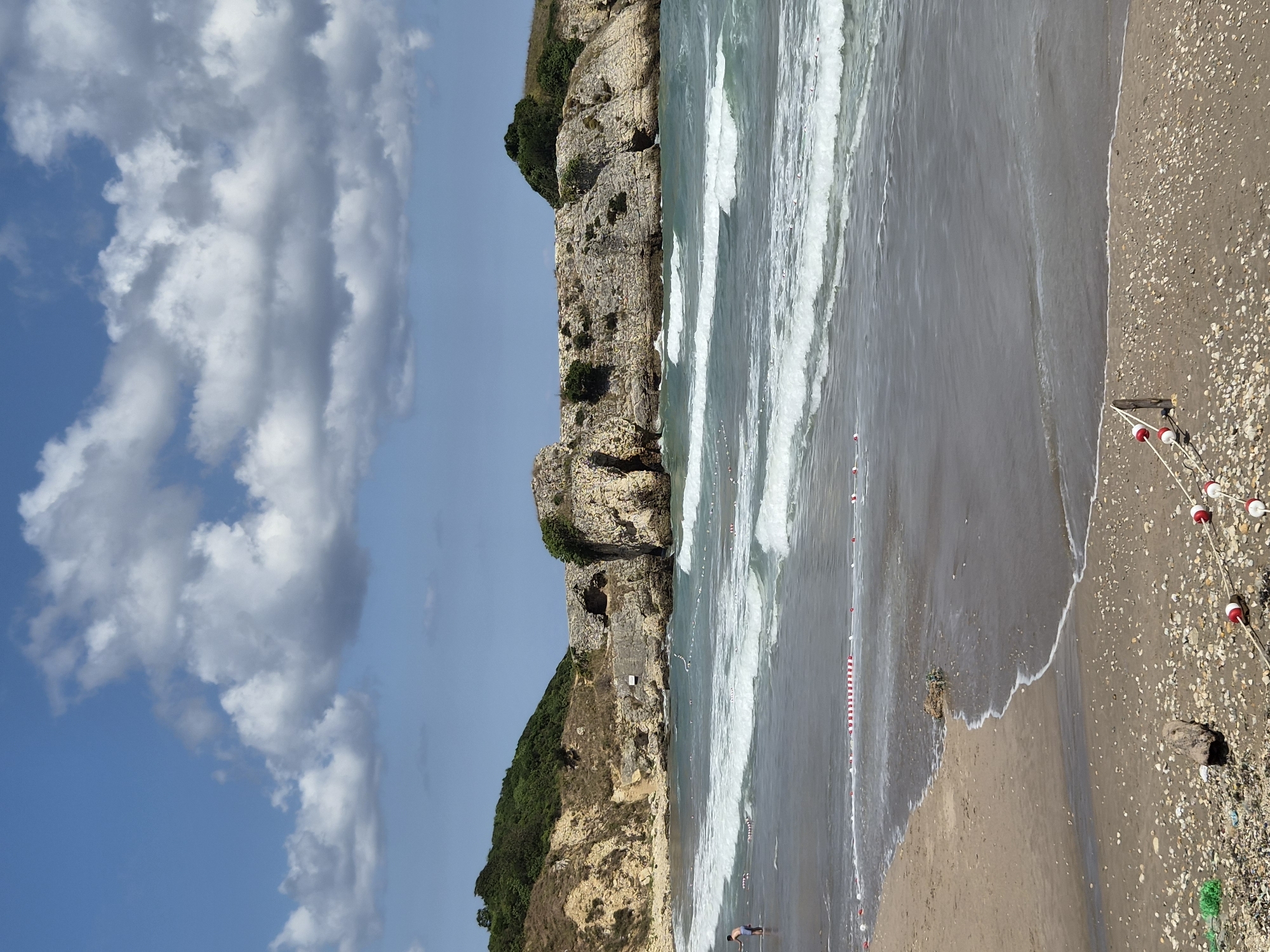 The image shows a view of the Black Sea coast at Şile, Turkey. In the foreground, a sandy beach is visible, strewn with shells and a piece of driftwood. A rope with red and white buoys runs along the edge of the sand. Further out, the sea is choppy with white-capped waves. In the middle ground, a person is standing in the shallow water. A floating rope demarcated by red and white buoys stretches across the water. In the background, rocky cliffs covered in green vegetation rise from the sea. The sky is blue with scattered white clouds.