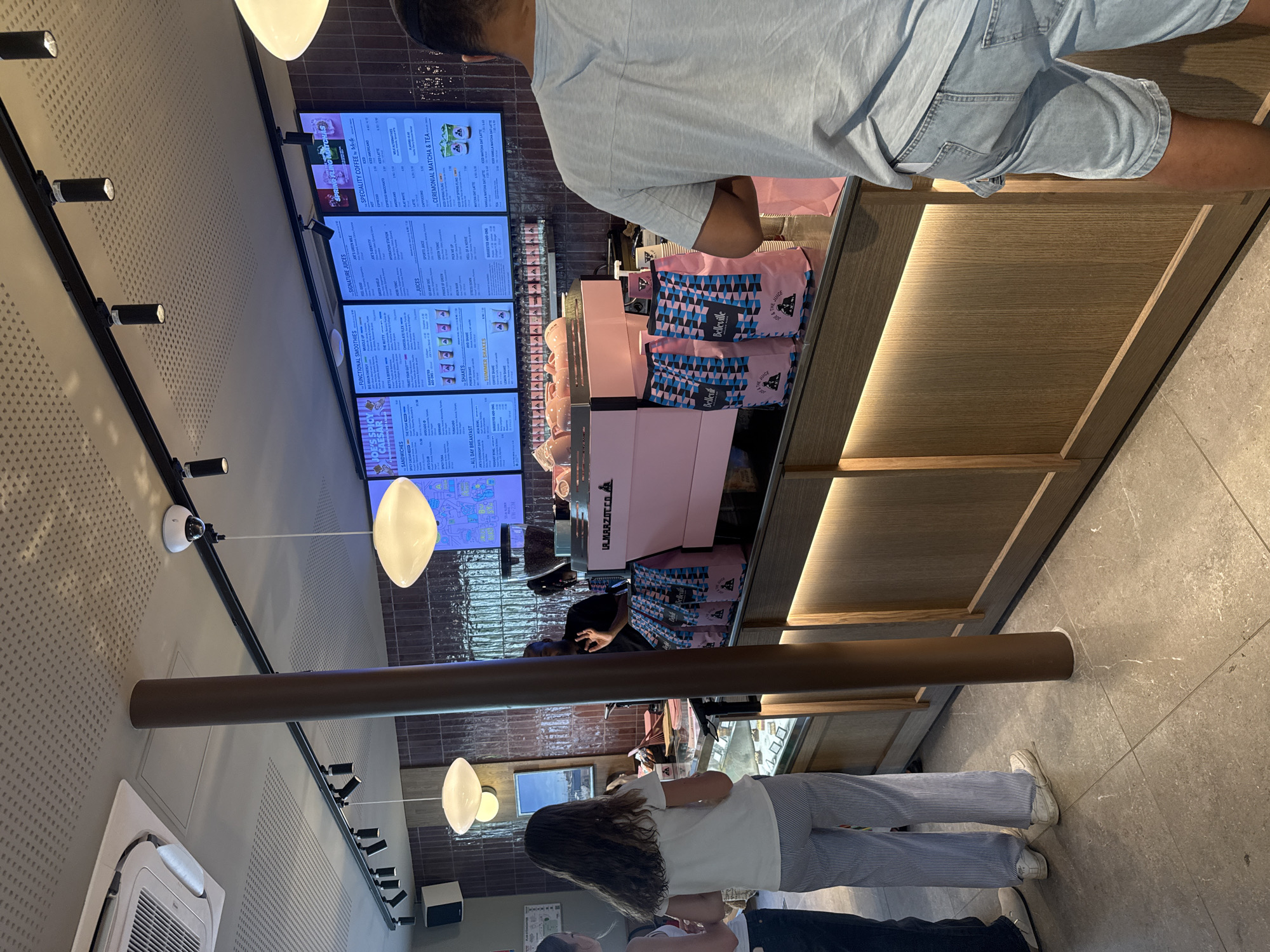 The image depicts the interior of a coffee shop, possibly a chain like Joe & The Juice, located in Paris, France. A barista, a dark-skinned man wearing a black t-shirt, is visible behind the counter, gesturing with his right hand. He is standing next to a bright pink La Marzocco espresso machine. In front of the counter, two women are standing. The woman on the left, wearing a white tank top and dark pants, is mostly out of frame. The woman in the center, facing away from the camera, wears a white t-shirt and striped pants. In the foreground, a man wearing a light blue t-shirt and denim shorts stands with his back to the camera, seemingly waiting to order. The shop has a modern aesthetic, with tiled walls, a light-up counter, and ceiling lights. Menus are displayed on screens above the counter, featuring various drinks and food items. Several pink and blue coffee bags are stacked on the counter. The floor is tiled in a neutral grey.
