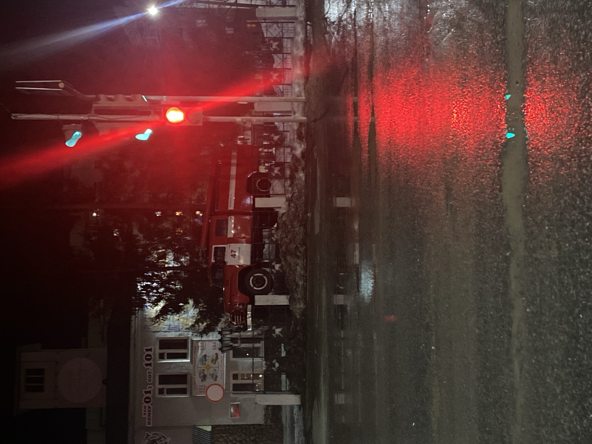 Here's a description of the image:
The photo, taken at night, shows a rainy street scene in the city of Chaikovsky, Russia, focusing on the reflection in a wet surface.
A fire engine is parked near a building with Russian text and the number "101" on it. The building's facade also displays several posters or advertisements. A traffic light displays red and green indicators, reflecting in the wet street. The overall lighting is poor, creating a dark and moody scene.