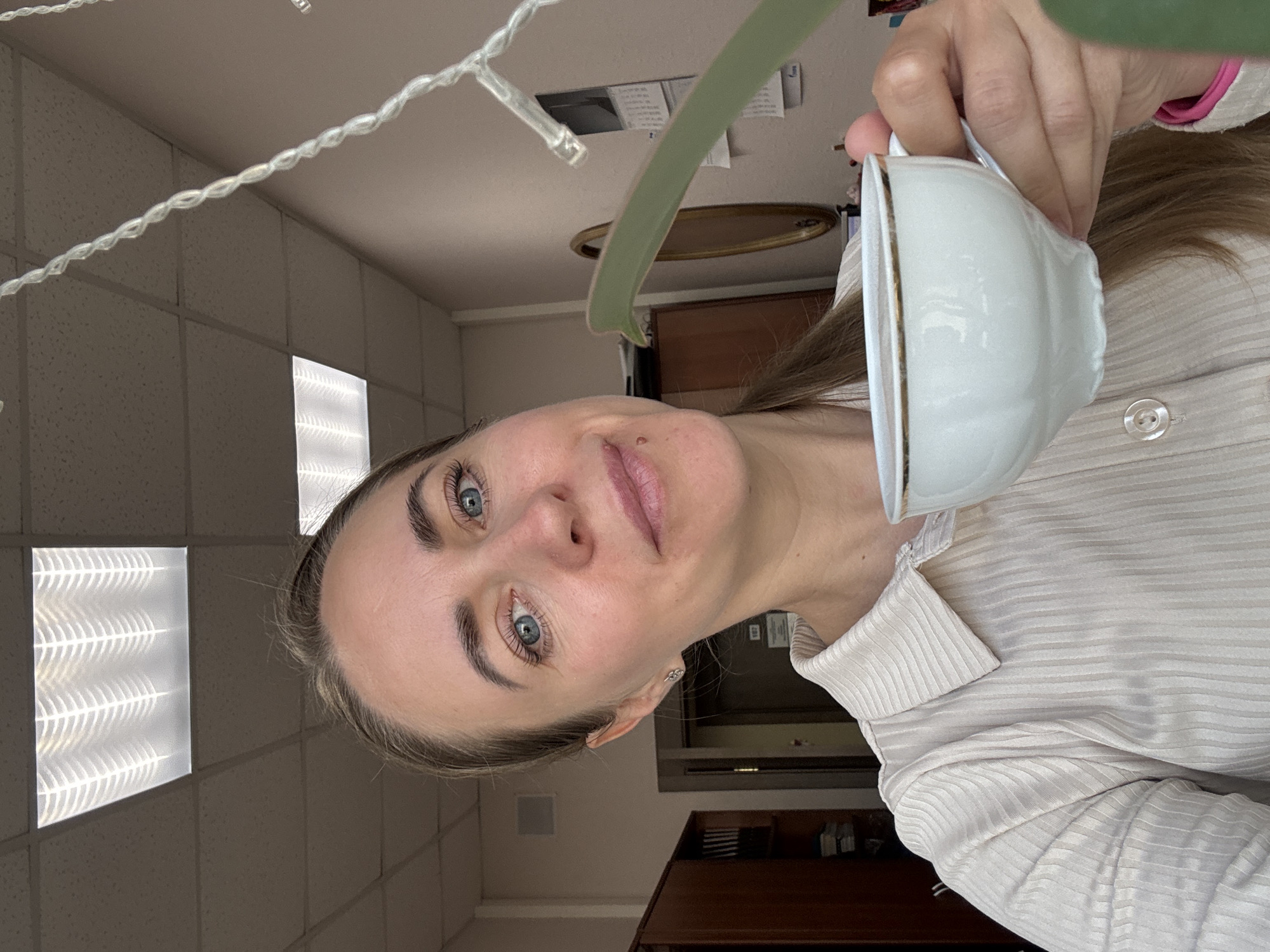 A woman is holding a teacup close to the camera, partially obscuring the background. The background includes a tiled ceiling with recessed lighting, a string of lights, a green leaf, and some furniture and cabinetry. The scene appears to be indoors. The location of the city of Chaikovsky, Russia, is not discernible from this image.