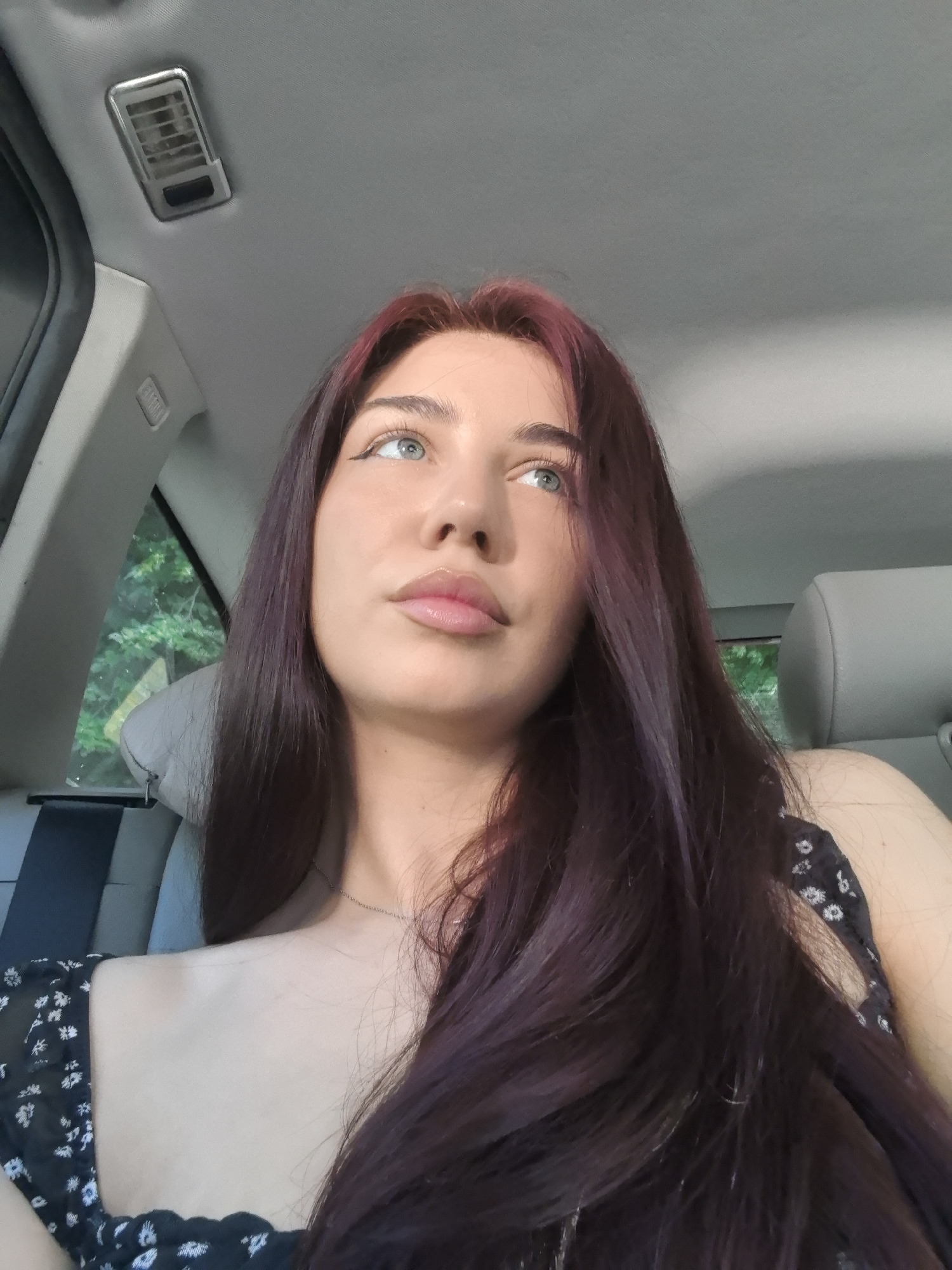 The image features a young woman inside a car during the daytime, depicted in a close-up portrait from the chest up. She has long, dark hair with distinct reddish-purple highlights, particularly visible near her roots. Her light blue-green eyes are accentuated with winged eyeliner, and she looks upwards and slightly to the right with a pensive or contemplative expression. Her lips are full and appear glossy. She is wearing a dark-colored top with a white floral pattern and a thin silver chain necklace around her neck.
The setting is the interior of a car, indicated by the grey headliner, an overhead air vent on the ceiling, and the design of the car seats. Through the window on the left side, lush green foliage is visible, suggesting an outdoor environment with trees or bushes. A blurry yellow triangular shape, possibly a road sign, is faintly discernible within the greenery. The bright, natural lighting indicates it is daytime. No specific activities are ongoing beyond her sitting and looking out. There is no visible text in the image. The location context provided is Stara Zagora, Bulgaria.