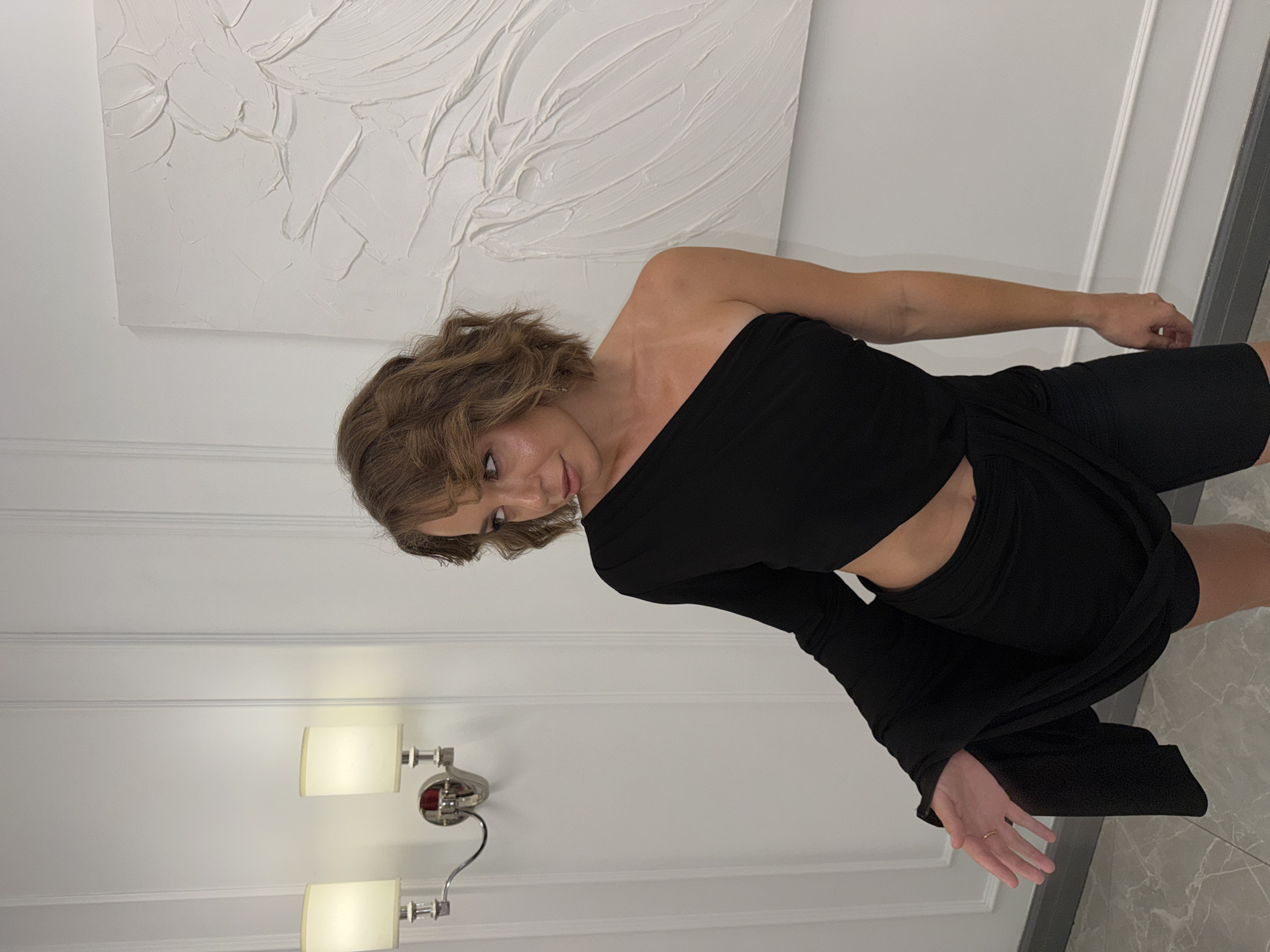 In Moscow, Russia, a woman stands in a room with white walls featuring vertical paneling and ornate molding. A contemporary abstract artwork in white hangs on the wall behind her. To her left, a modern double wall sconce with cream-colored shades illuminates the space. The woman, with wavy brown hair, is wearing a black one-shoulder dress with a cut-out detail at the waist and a flowing skirt. She is looking to her left, with her left arm extended. Her pose suggests movement, perhaps mid-twirl. The flooring is a polished grey marble.