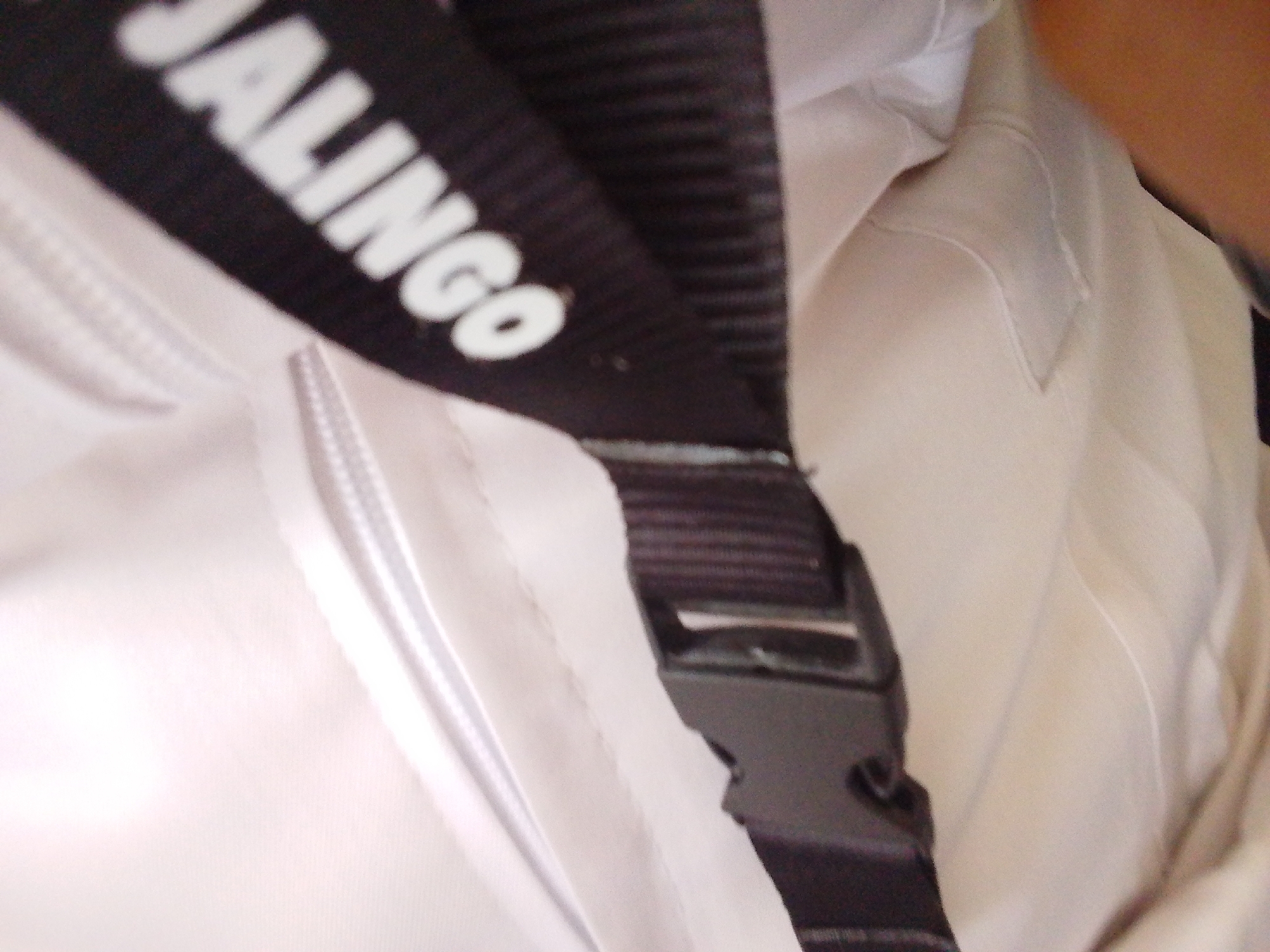 The image shows a close-up view of a black strap with white lettering that reads "JALINGO". The strap is attached to a black plastic buckle. The strap and buckle are resting on a white fabric, possibly part of a garment or bag. The background is out of focus, but appears to be a light-colored surface. There is no indication of the location in Kuson, Nigeria.