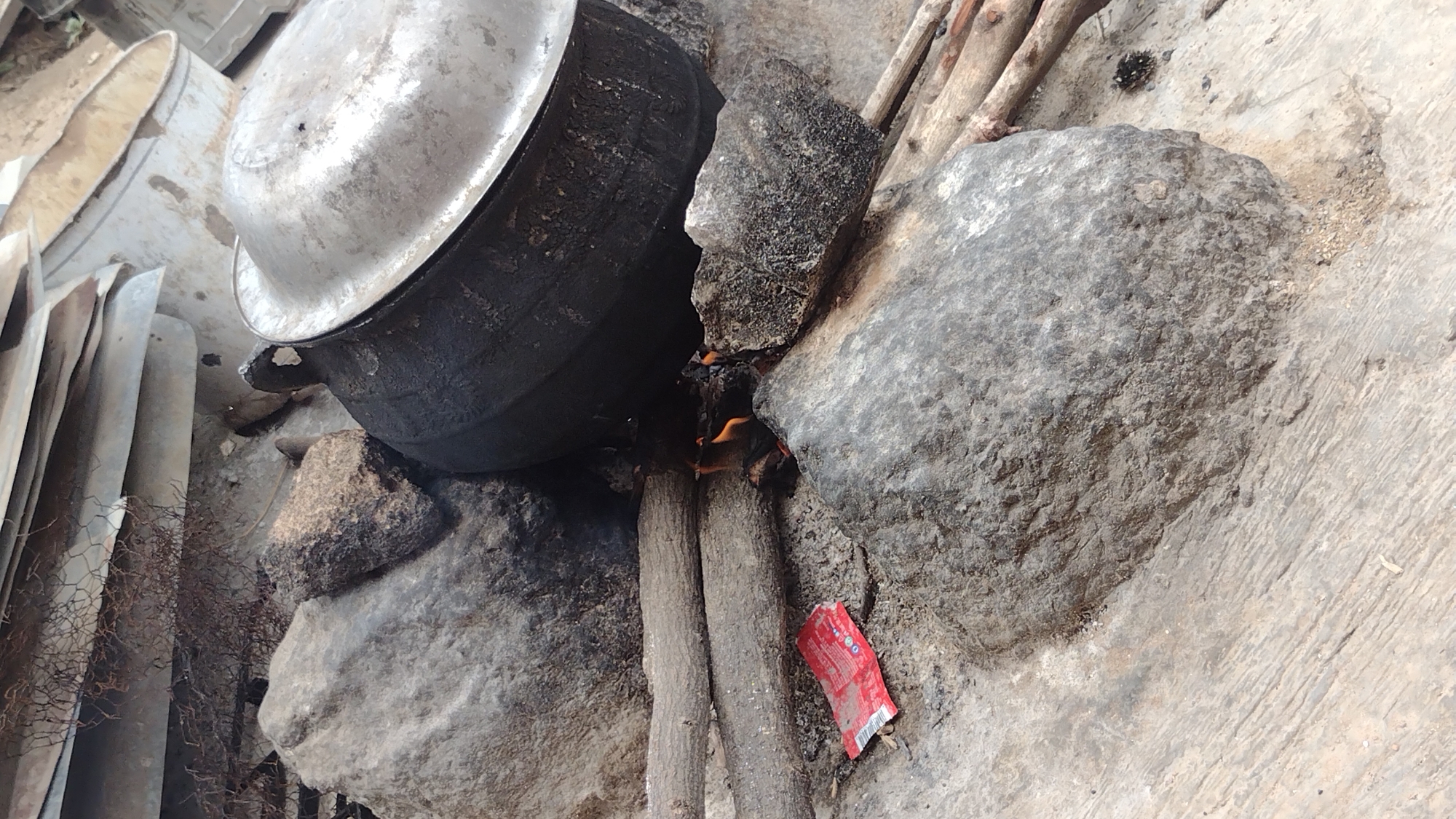 This image depicts a rustic outdoor cooking setup, likely located in Jos, Nigeria, given the provided context. A large, dark-colored cooking pot with a reflective silver lid is positioned over an open fire. The pot is supported by a makeshift arrangement of several substantial, irregularly shaped rocks and two thick wooden logs, which also serve as fuel for the fire. Bright orange flames are visible emanating from between the logs, indicating active cooking.
The ground surrounding the fire pit is a mix of rough earth and what appears to be a light-colored concrete or paved surface, particularly towards the right side of the frame. In the background on the left, several pieces of corrugated metal sheets are stacked, alongside some rusted wire mesh. On the ground near the fire, nestled between a log and a rock, lies a small, crumpled red wrapper with white text, possibly from a food item. Additional wooden branches, presumably more fuel, are visible in the upper right portion of the image.
The scene is brightly lit, suggesting it is daytime. No people are visible in the photograph, but the setup clearly indicates an ongoing or recently used outdoor kitchen for preparing food. The overall impression is one of a functional, basic cooking arrangement commonly found in rural or informal urban settings.