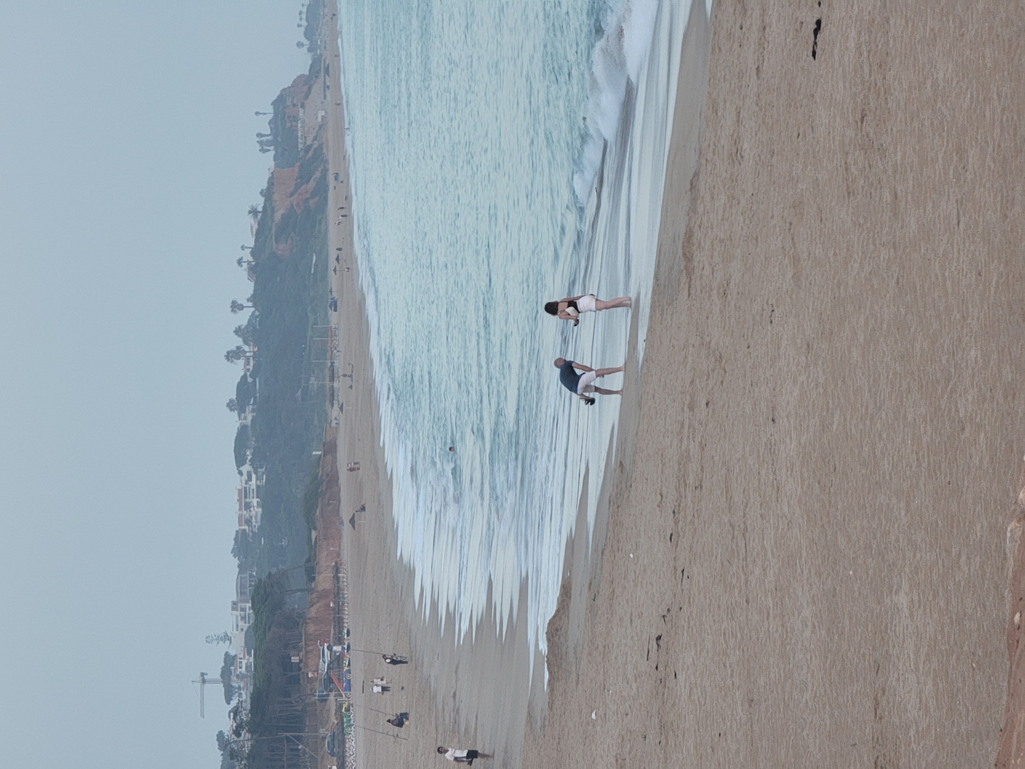 The image depicts a wide, sandy beach with the ocean in the background, likely in Quarteira, Portugal, given the context. In the foreground, two individuals are standing near the water's edge. One is a person wearing a dark shirt and white shorts, bending over with their hands near the sand. The other is a person wearing a white bikini top and white shorts, facing the first individual. A few feet away in the water, another person is visible swimming. The tide is coming in, with waves gently breaking on the shore.
Further up the beach, several other people are scattered, some appearing to be fishing with long poles. In the distance, a resort-like area with buildings and trees is situated on a cliff overlooking the beach. A construction crane is also visible on the horizon, suggesting ongoing development. The sky is overcast, creating a muted, diffused light. The overall scene is one of a somewhat quiet, possibly overcast day at the beach with recreational activities taking place.