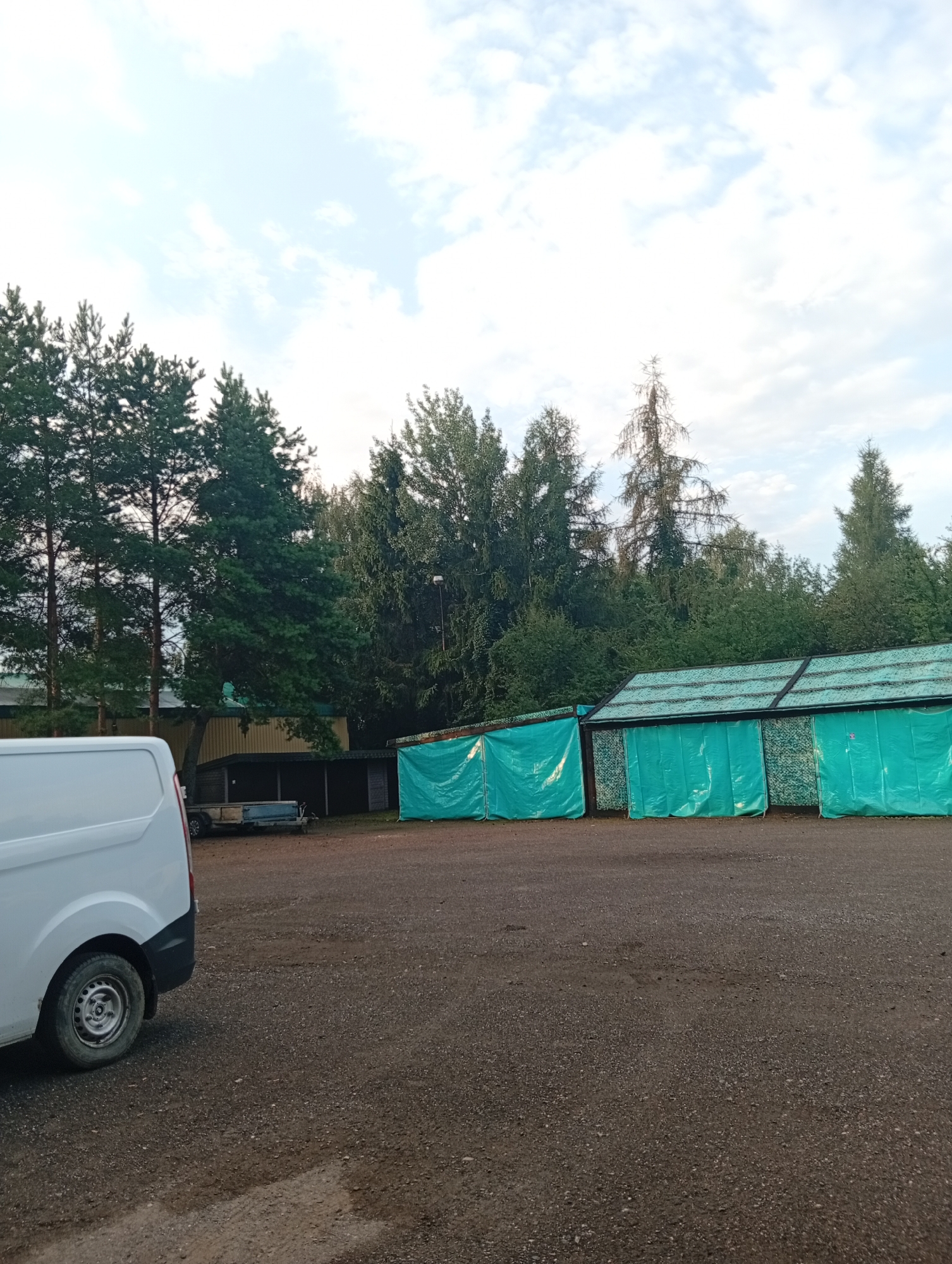 The image depicts an outdoor scene with a gravel surface in the foreground. On the left, the rear portion of a white van is visible. Behind the van and to the right, there are several structures. One structure appears to be a shed with an open front, and next to it are three covered stalls or bays with turquoise tarpaulins. These stalls are lined up in a row and have a slatted roof structure. In the background, a dense line of trees, primarily coniferous, forms a backdrop. The sky above is partly cloudy. A small, blue trailer is parked between the van and the shed-like structure. The overall impression is one of an outdoor storage or service area, possibly at the edge of a wooded area. The location is identified as Kożuszki-Parcel, Poland.